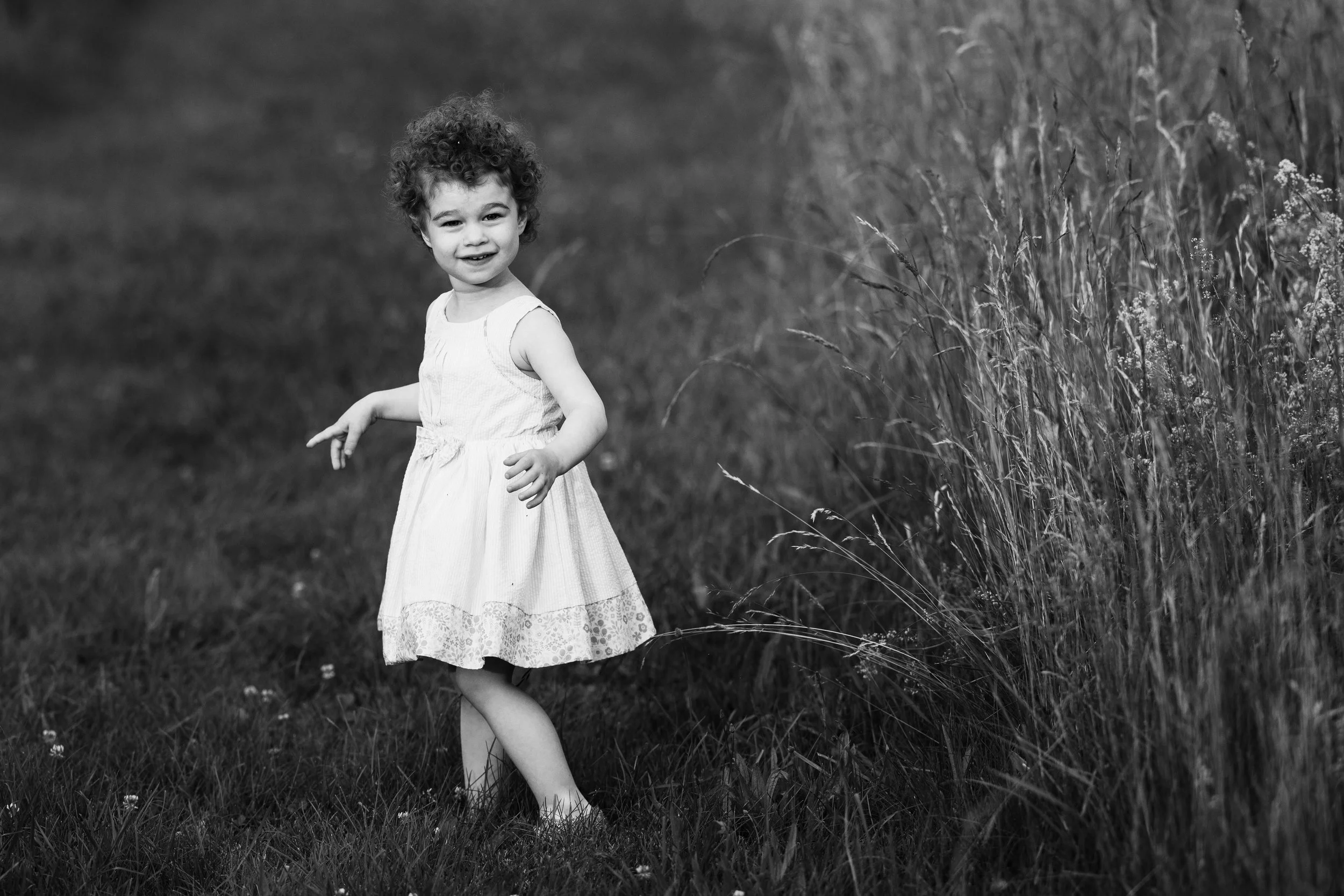 A young girl with curly hair wearing a sleeveless white dress standing in a grassy field next to tall grass, smiling at the camera.