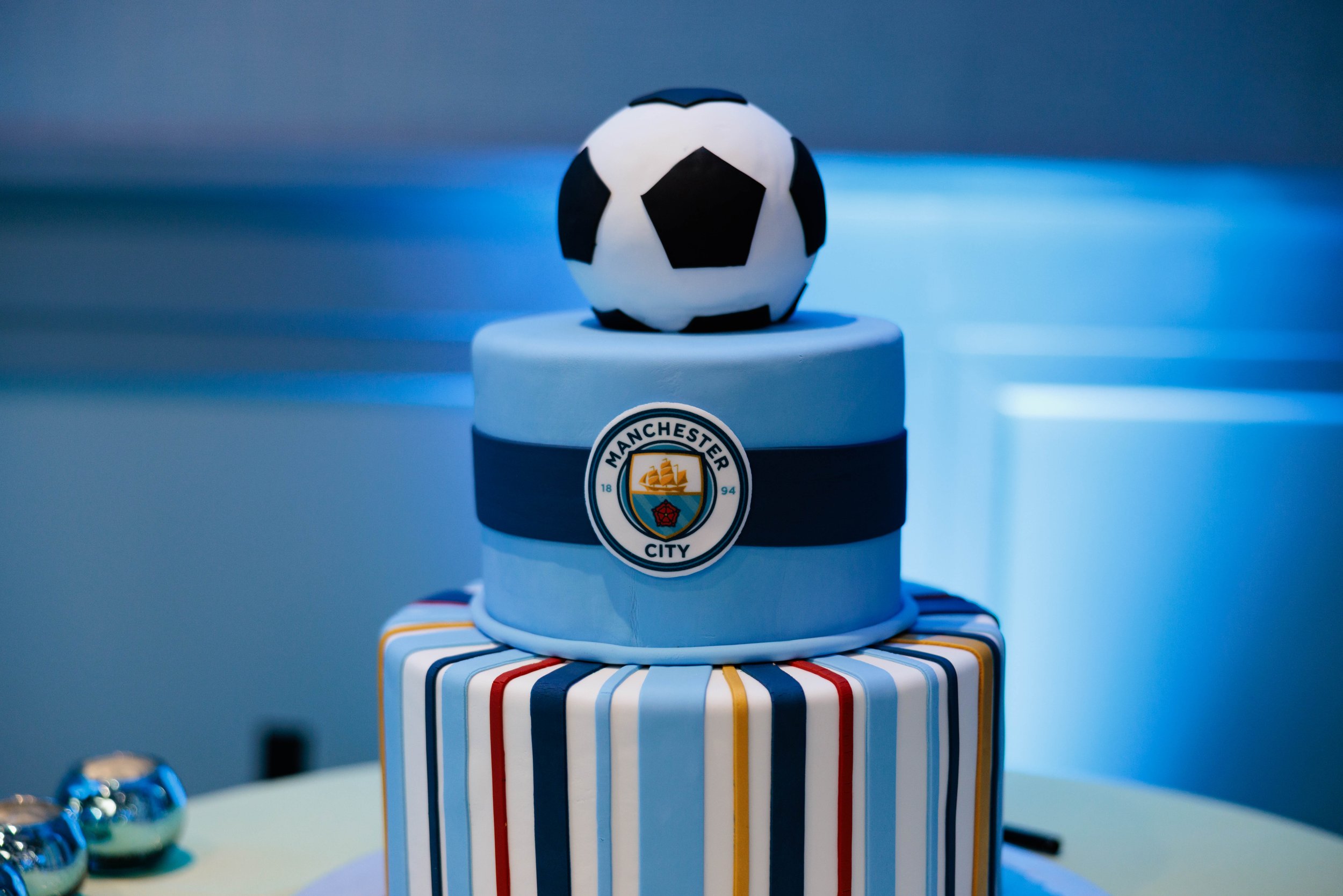A cake with two tiers featuring the Manchester City Football Club emblem on top, topped with a soccer ball decoration.