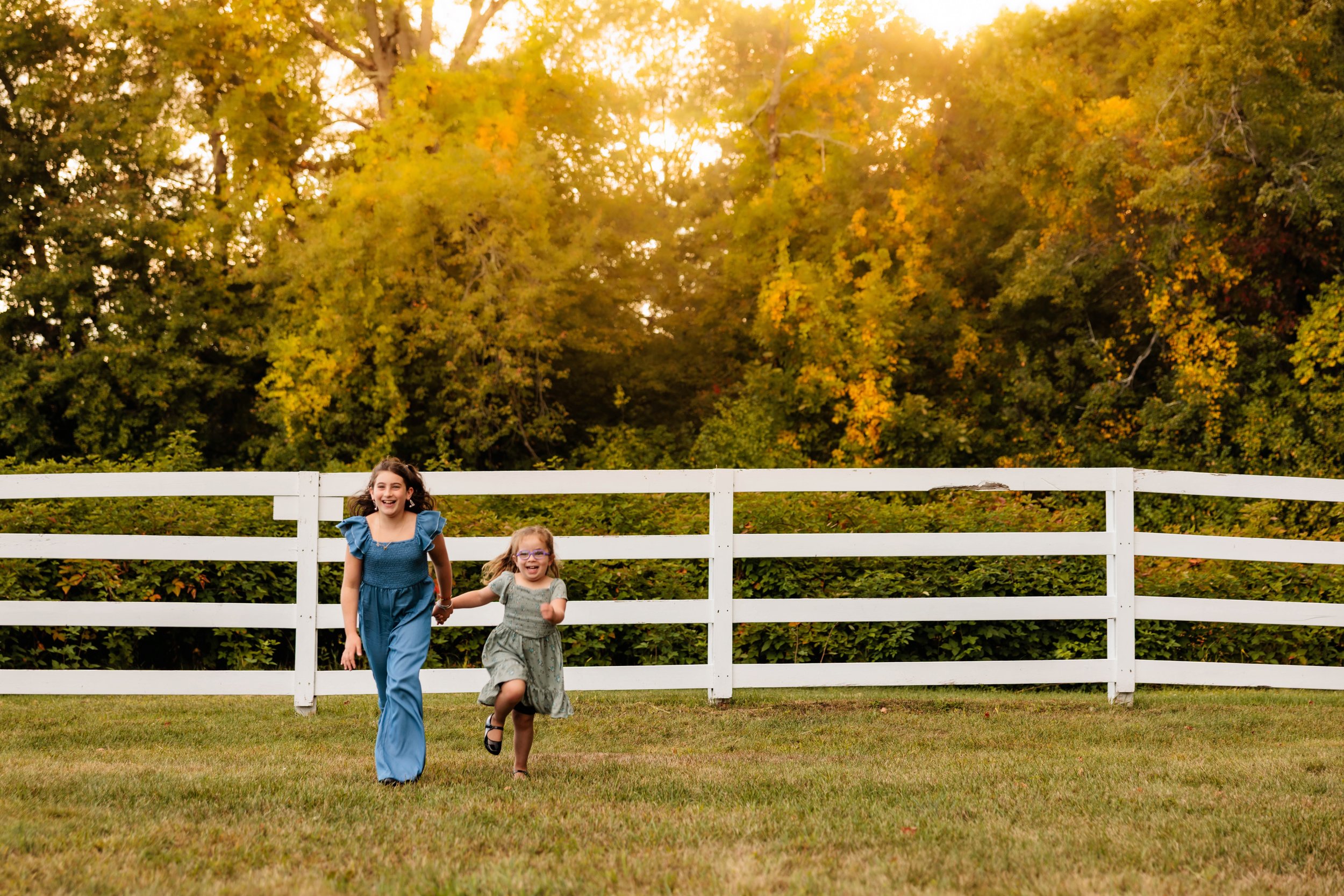 Two young girls with glasses happily running and holding hands in a grassy yard with a white fence and autumn-colored trees in the background.