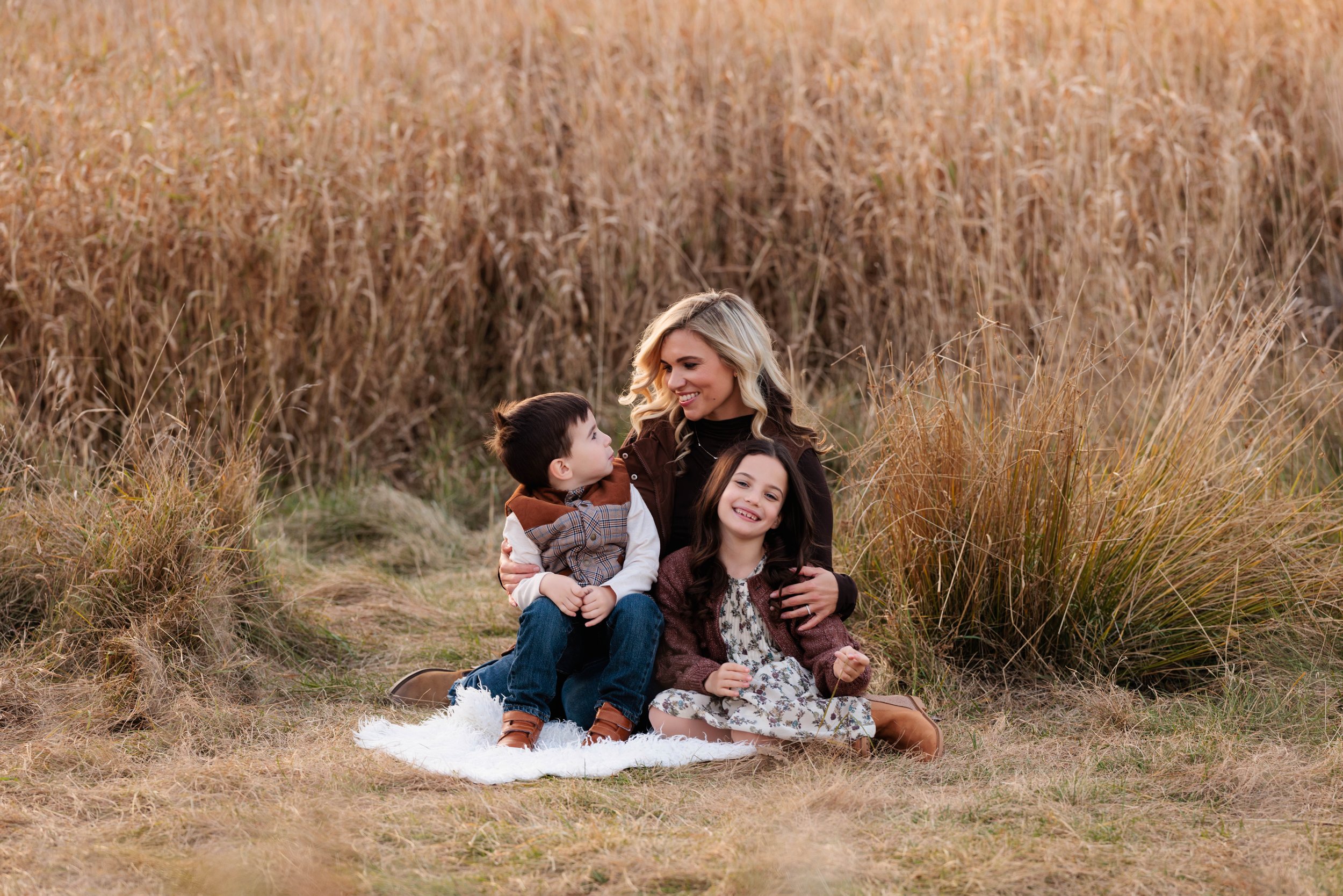 A woman with two young children sitting on a white blanket in a field of tall, golden grass during autumn. The children are smiling and looking at each other, while the woman is smiling warmly at them.