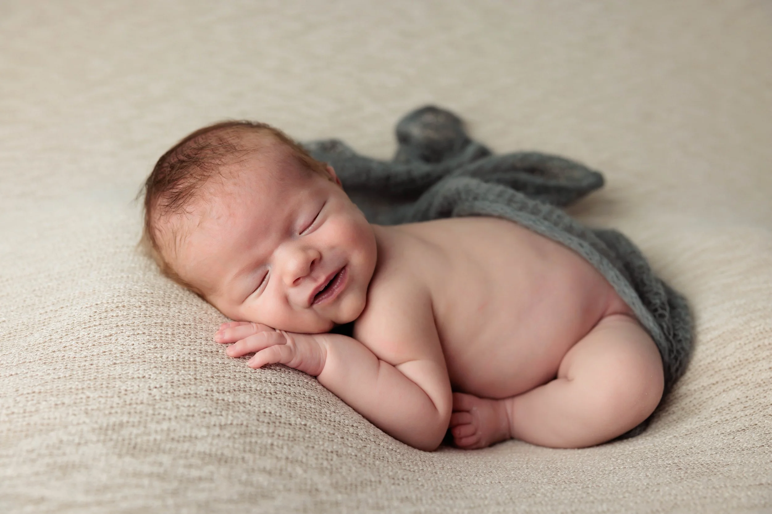 A smiling baby lying on a couch, resting its head on one hand, with a blanket partially covering its back.