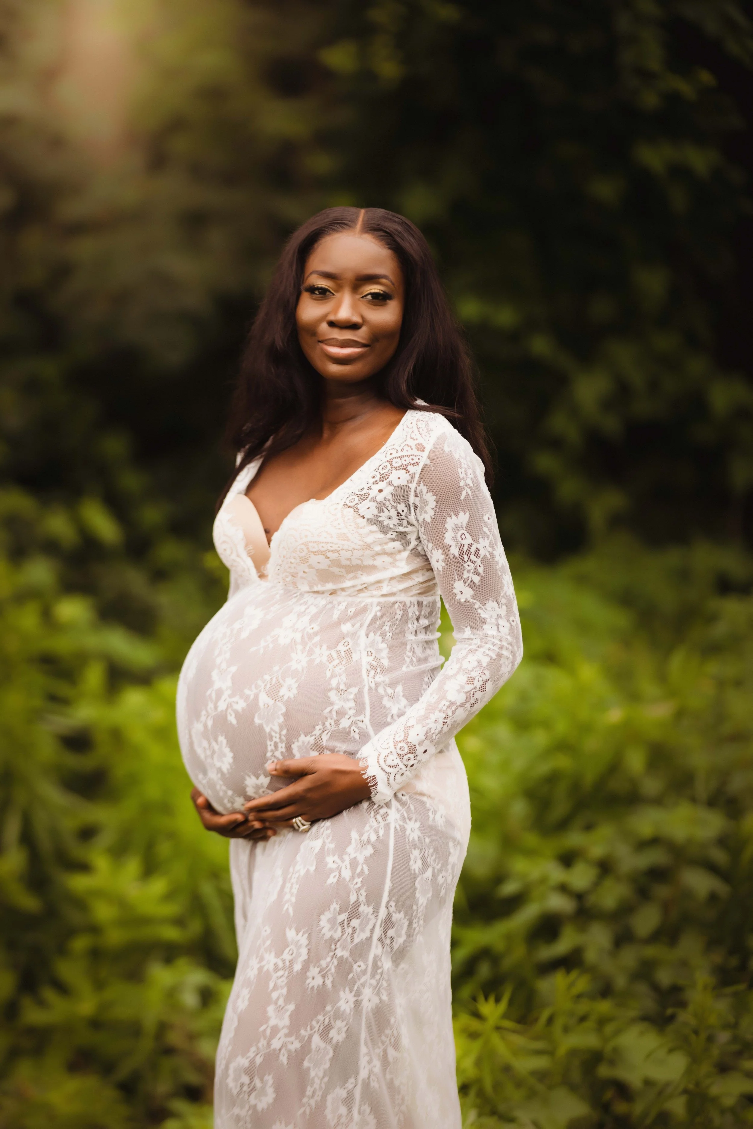 A pregnant woman in a white lace dress standing outdoors in a green, leafy setting, gently holding her baby bump.