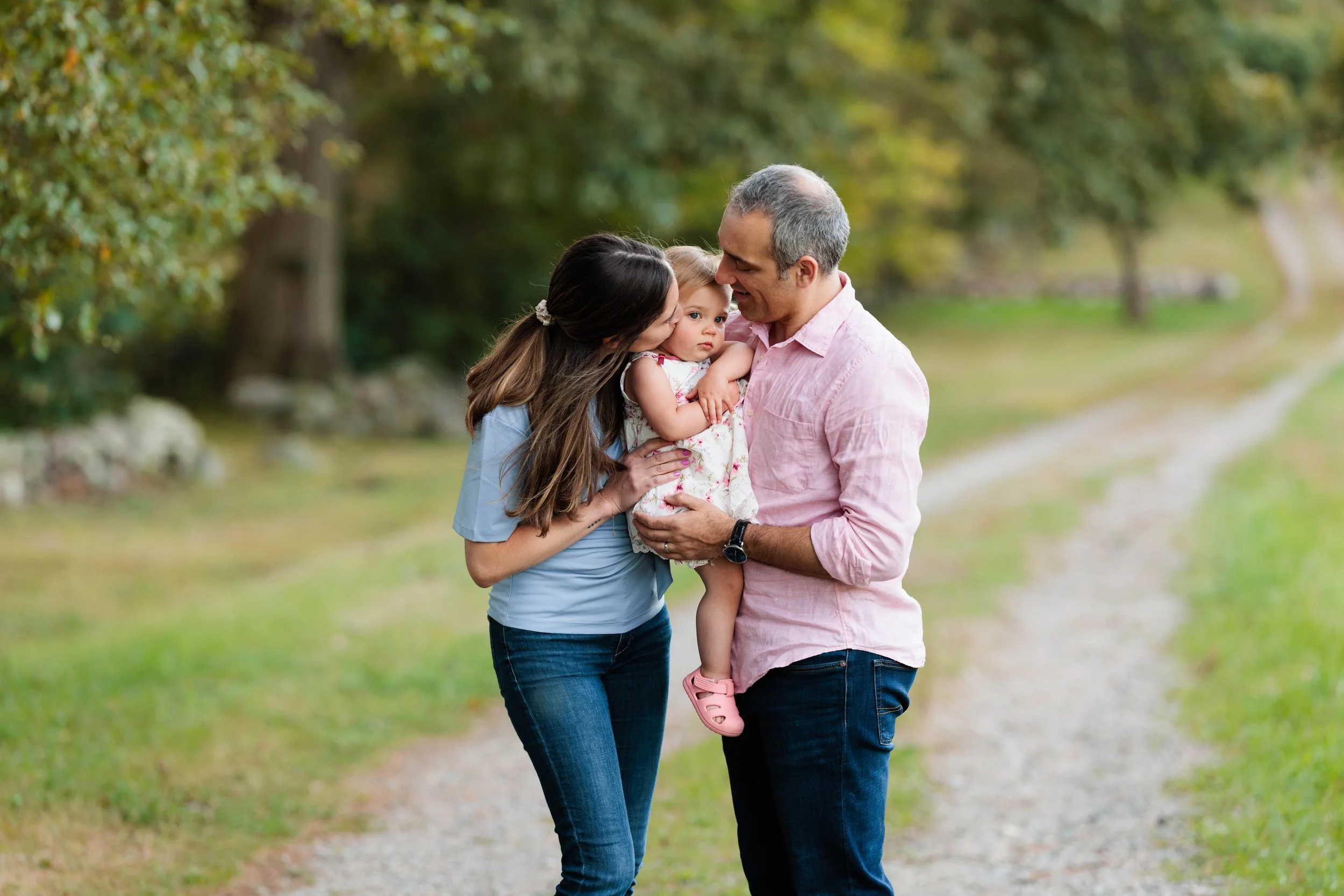 A family of three standing on a dirt path in a park, with trees and greenery in the background. The mother is leaning in to kiss the young girl, who is being held by the father. The girl has blonde hair and is wearing a floral dress and pink shoes.