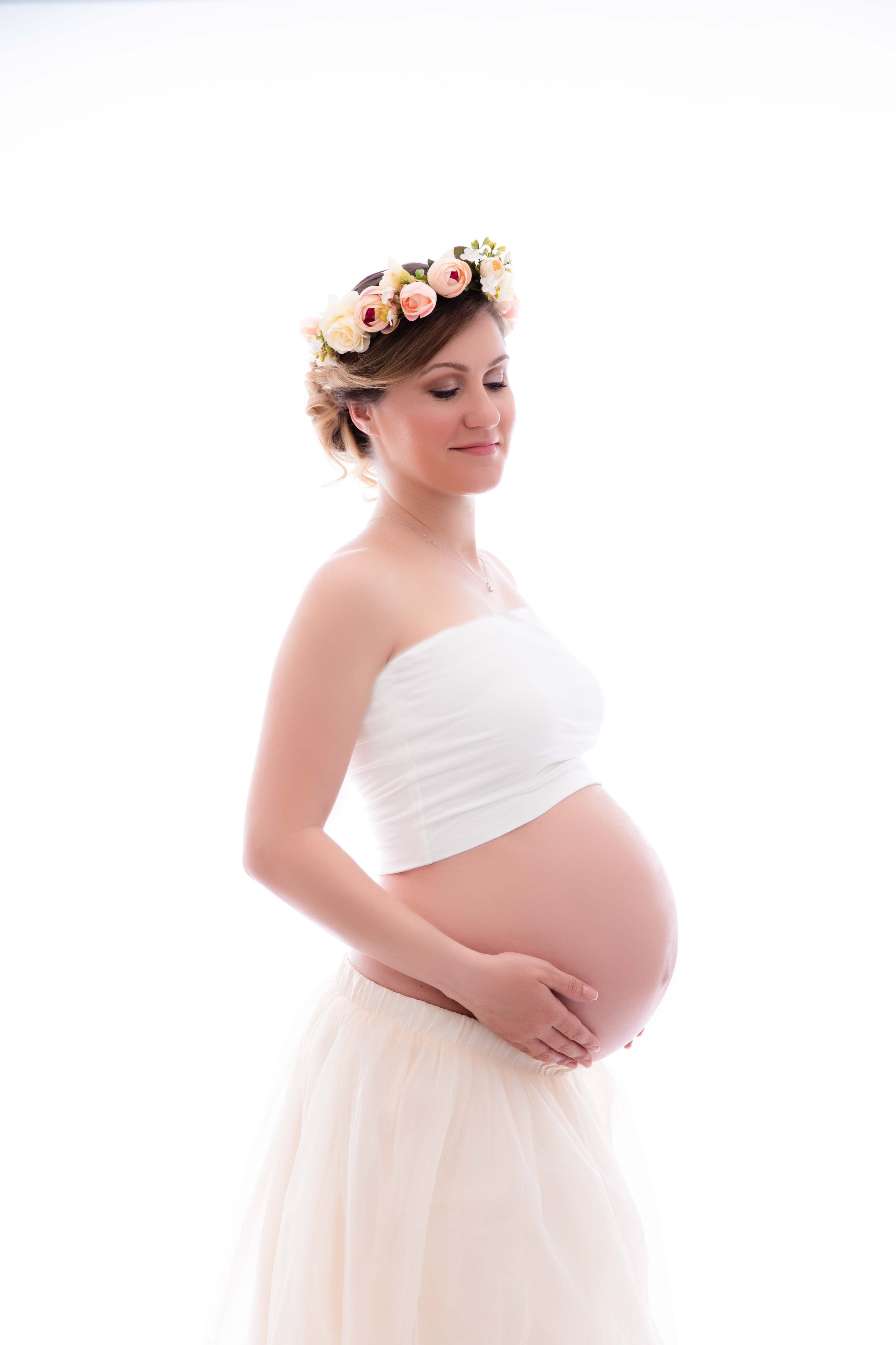 Pregnant woman with headflower, white crop top, and cream-colored skirt, gently holding her belly and standing against a bright background.