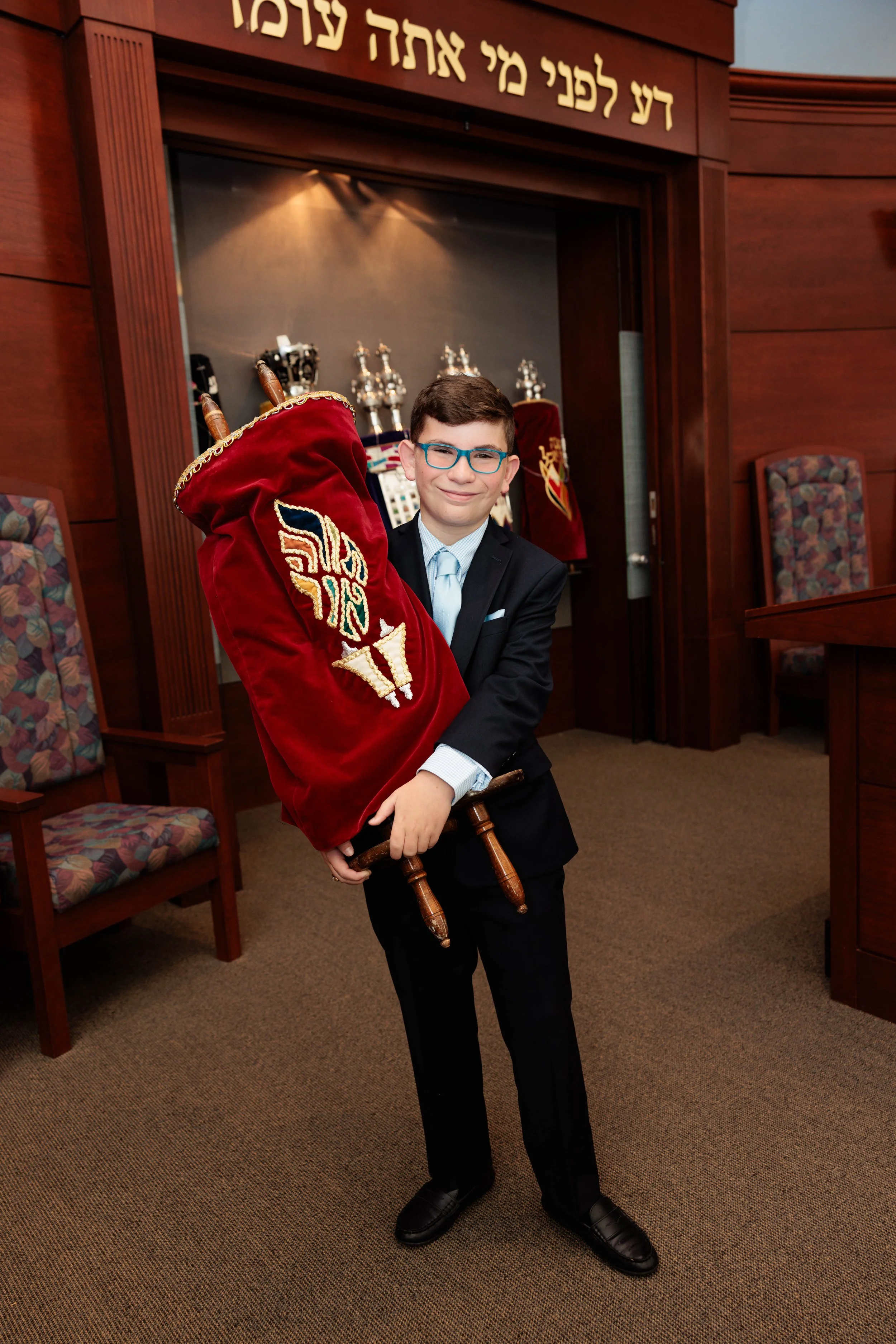 A young boy dressed in a suit holding a ceremonial Torah scroll in a synagogue.