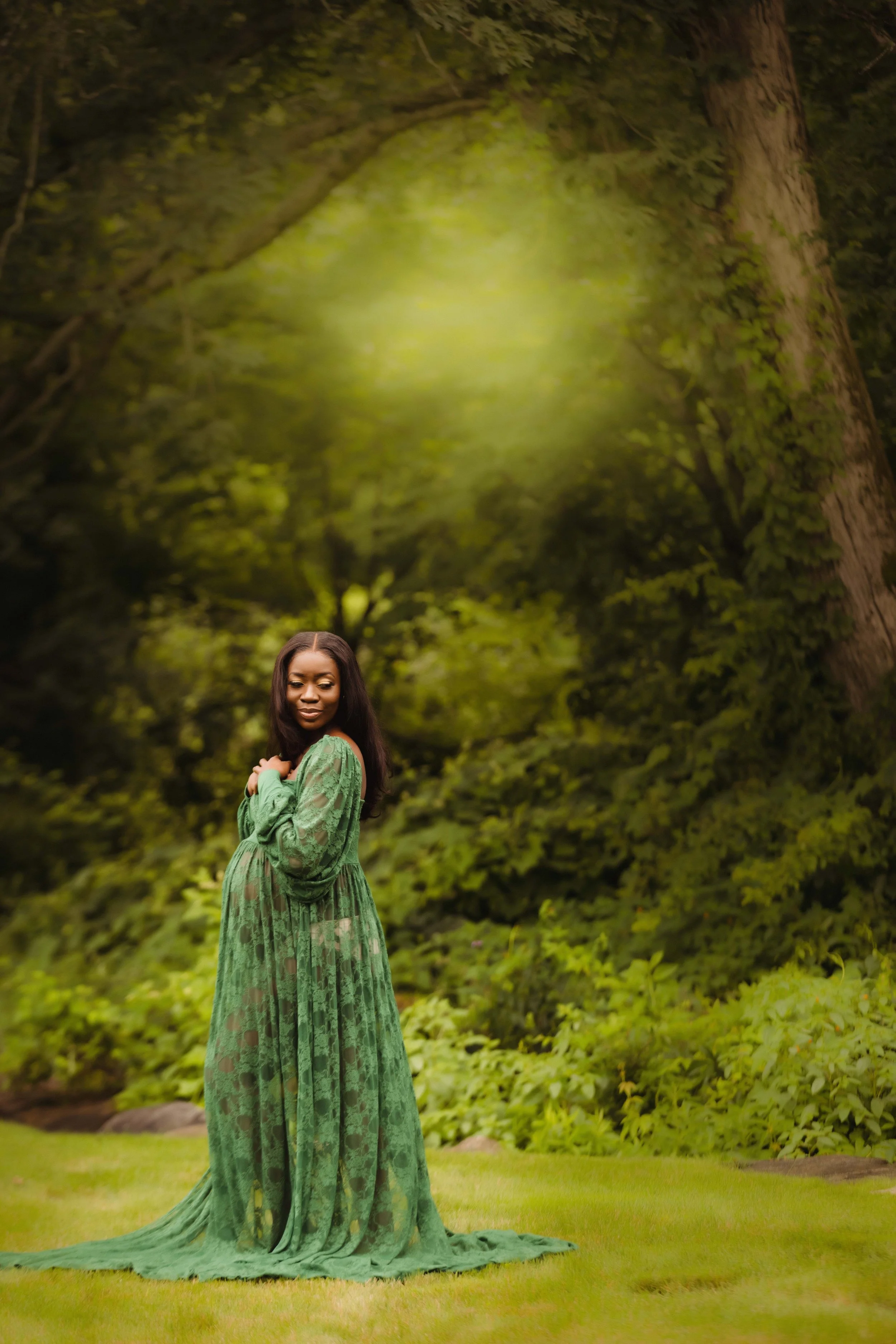 A woman in a flowing green dress standing on grass in front of a dense, leafy forest with a large tree trunk behind her.