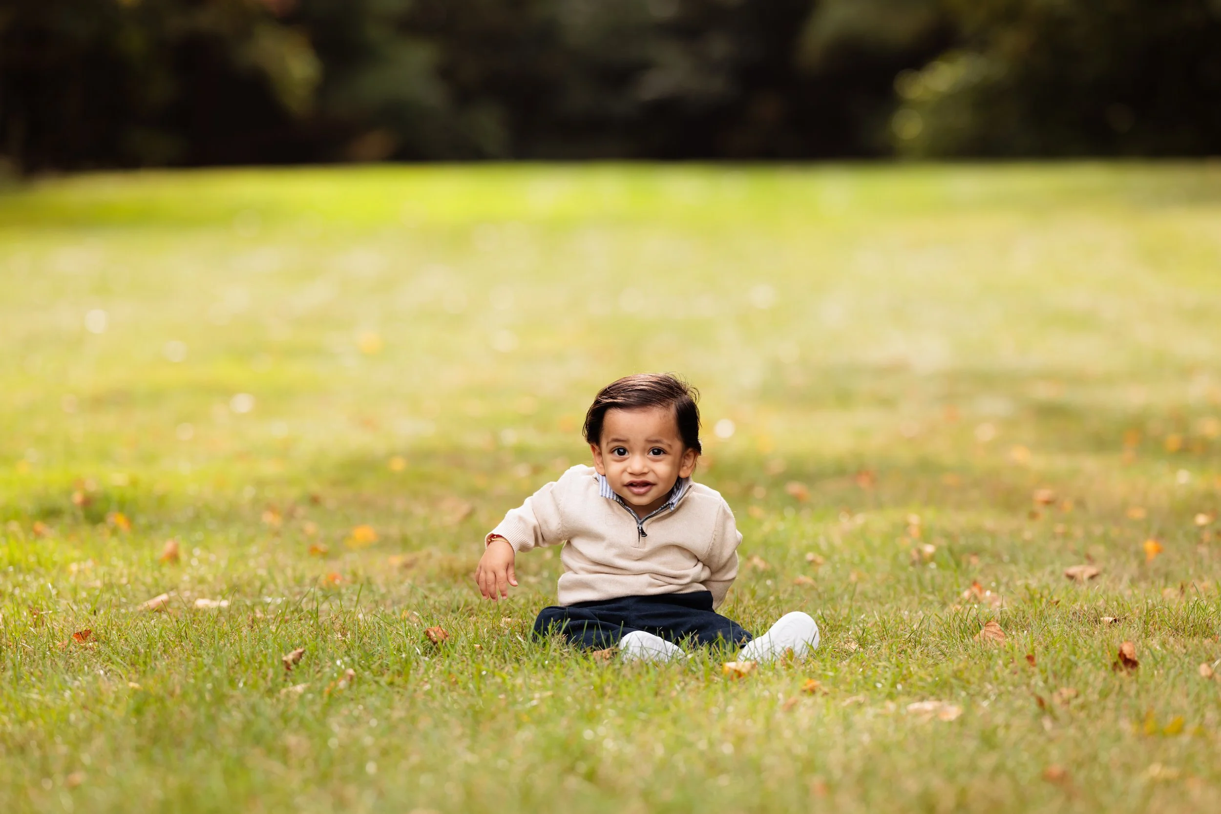 A smiling young boy sitting on the grass in a park, with trees in the background.