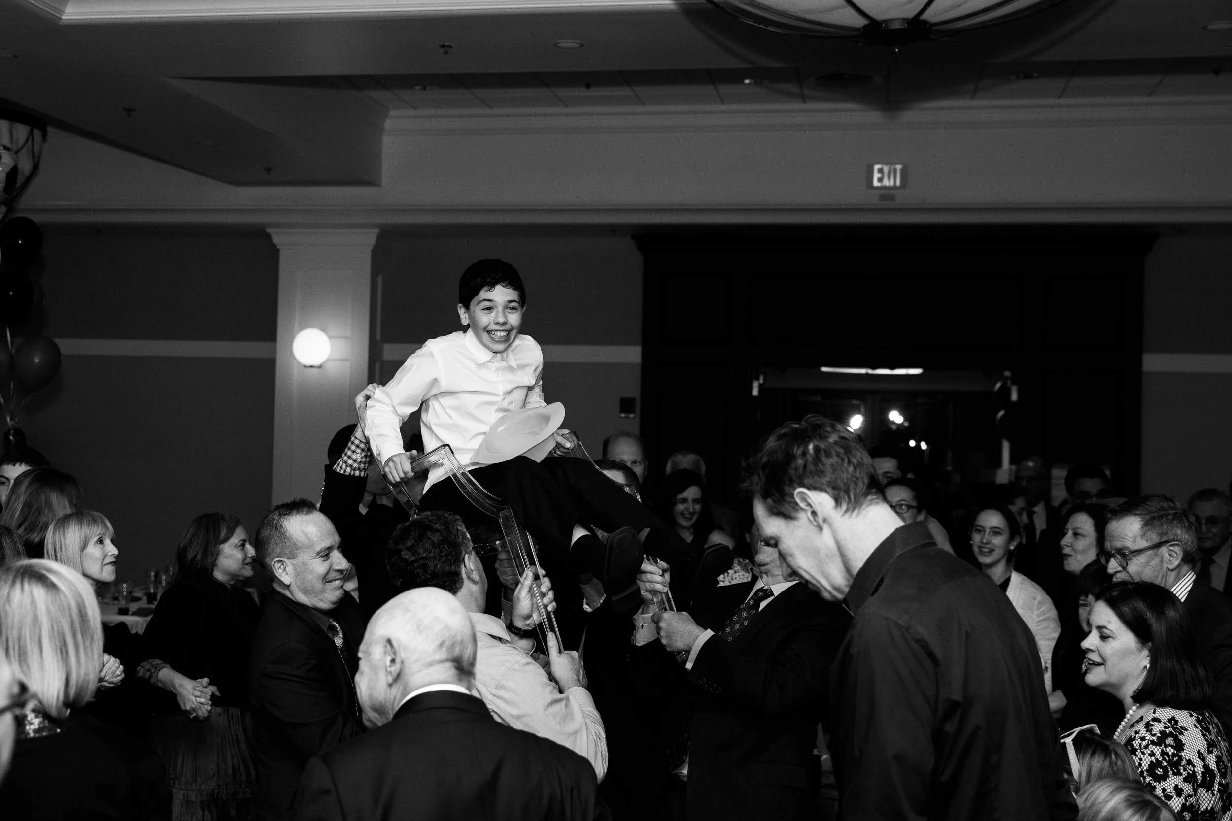 A young boy is lifted in the air on a chair by adults at a celebration or party, surrounded by smiling guests.