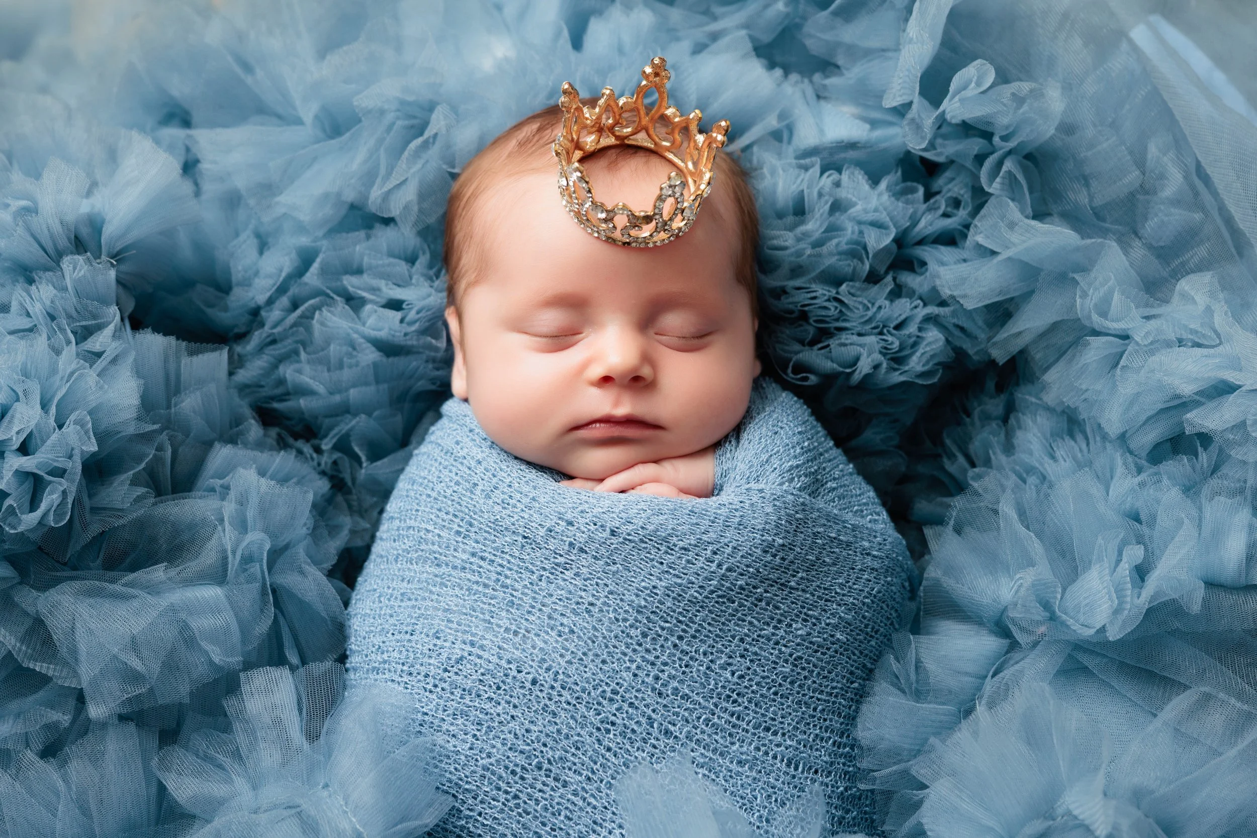 A newborn baby sleeping peacefully, wrapped in a blue blanket, with a small crown headband on their head, surrounded by blue ruffled fabric.