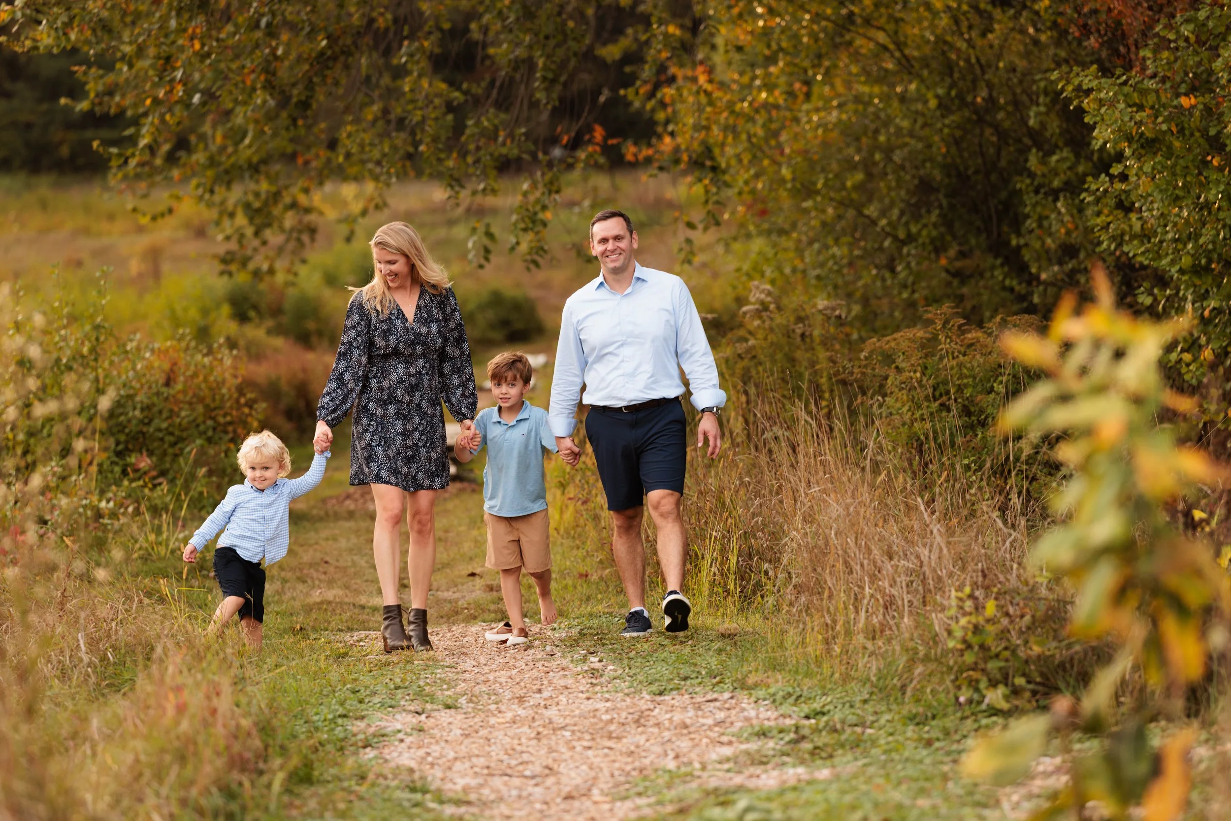 Family of four walking on a dirt path through a wooded area with autumn foliage, holding hands and smiling.