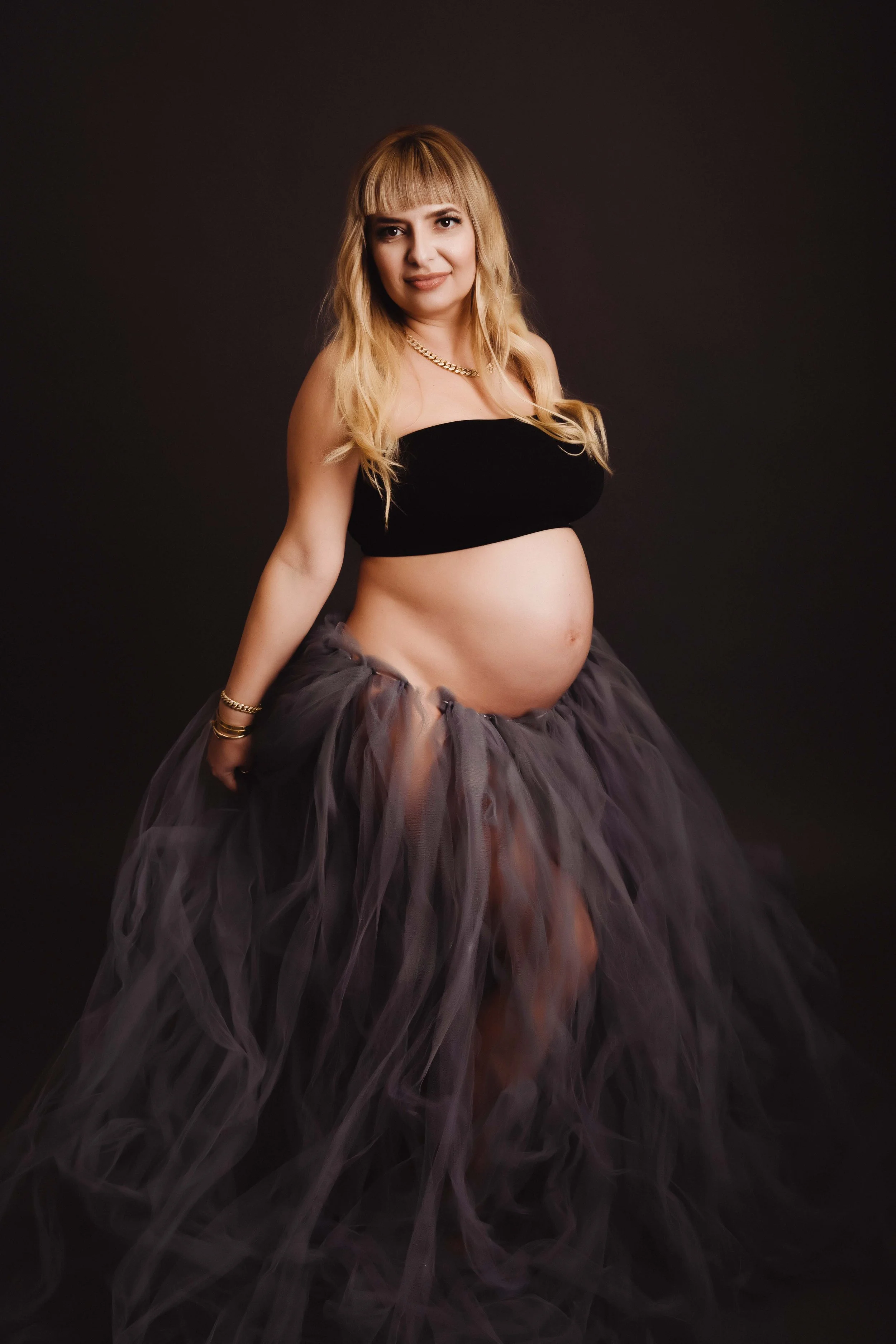 A pregnant woman with blonde hair wearing a black tube top and a flowing dark tulle skirt, standing against a dark background.
