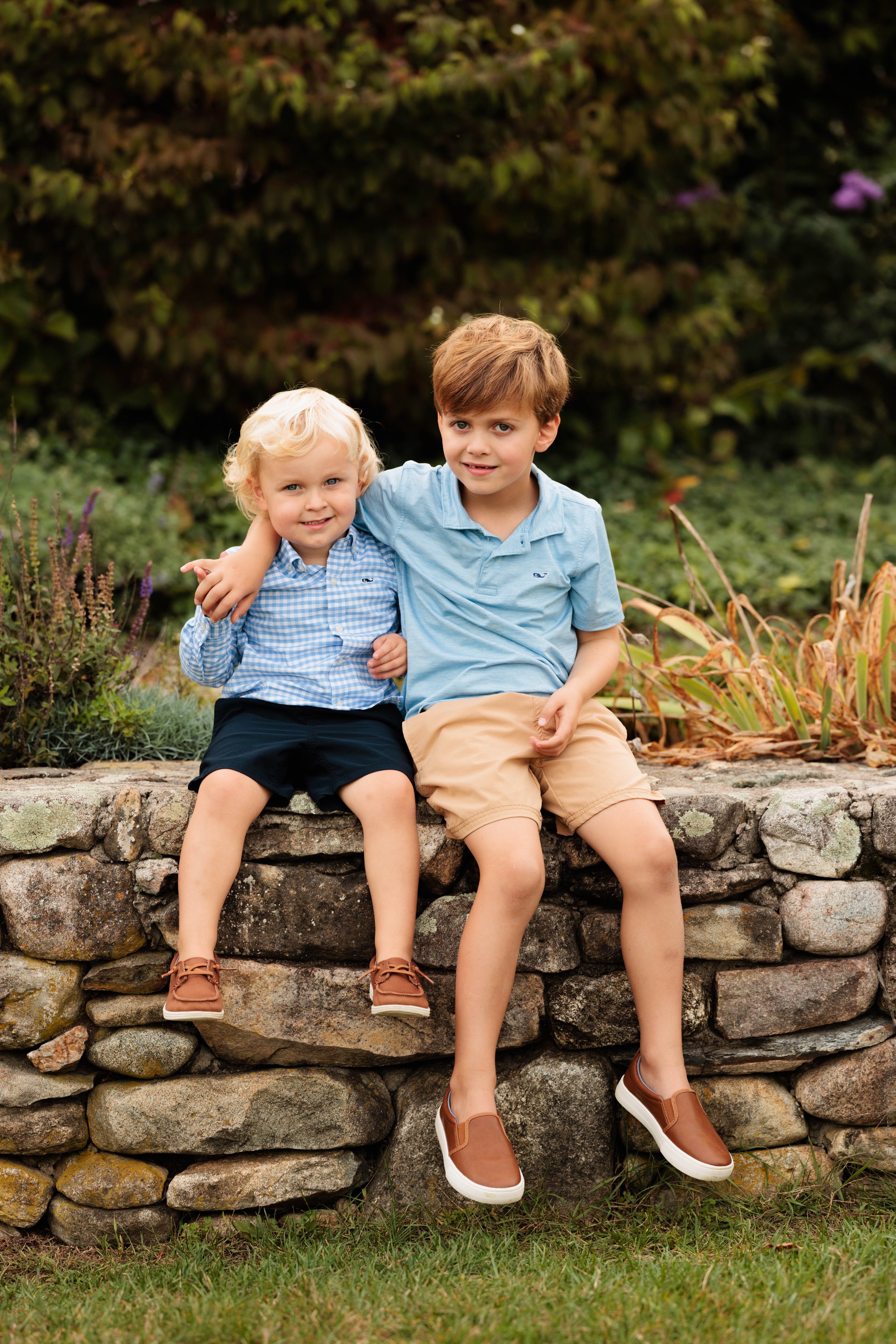 Two young boys sitting on a stone wall in a garden, smiling at the camera, with one boy' arm around the other.