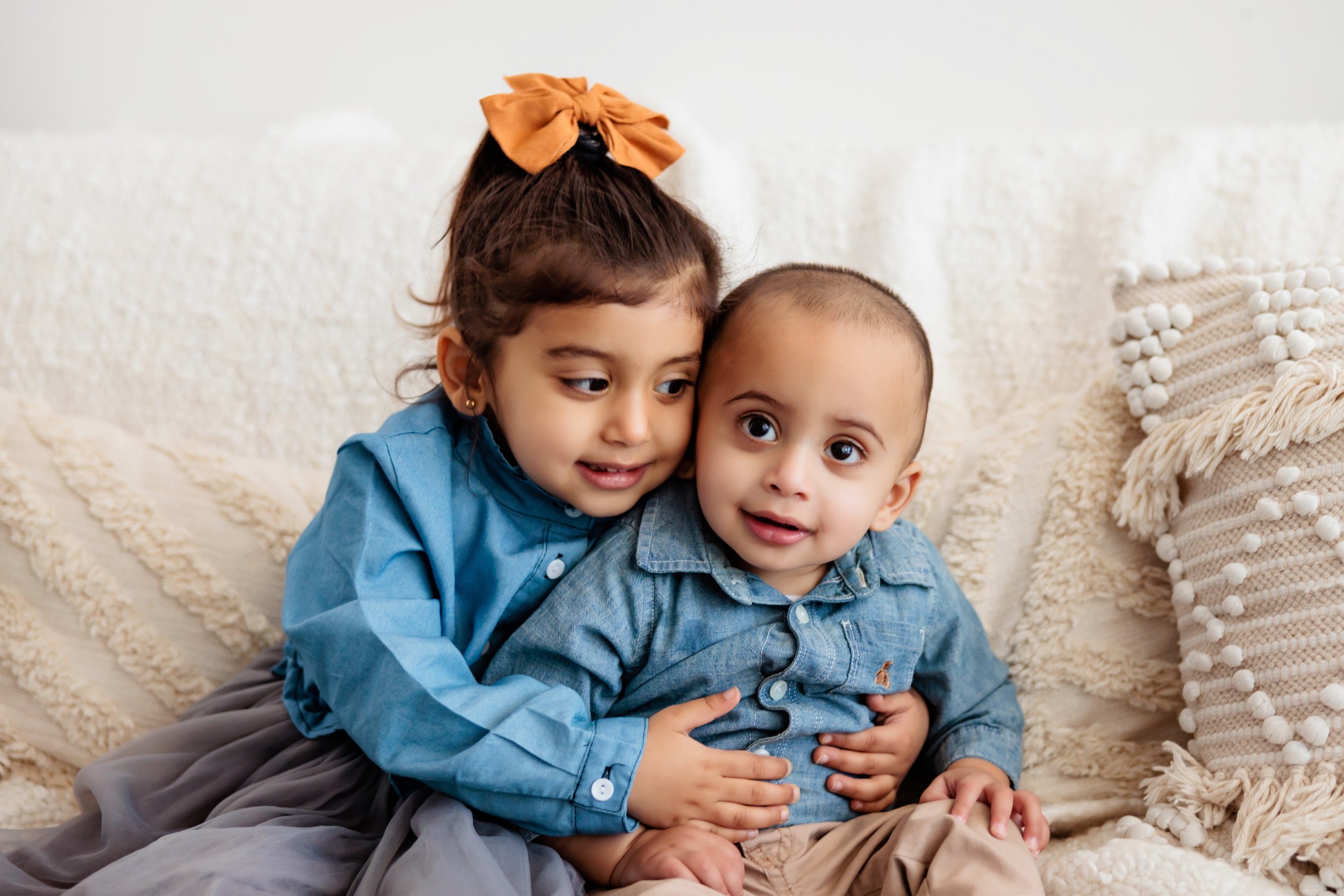 Two young children, a girl and a boy, sitting close together on a white couch with textured pillows, wearing denim and blue shirts, embracing and looking at the camera.