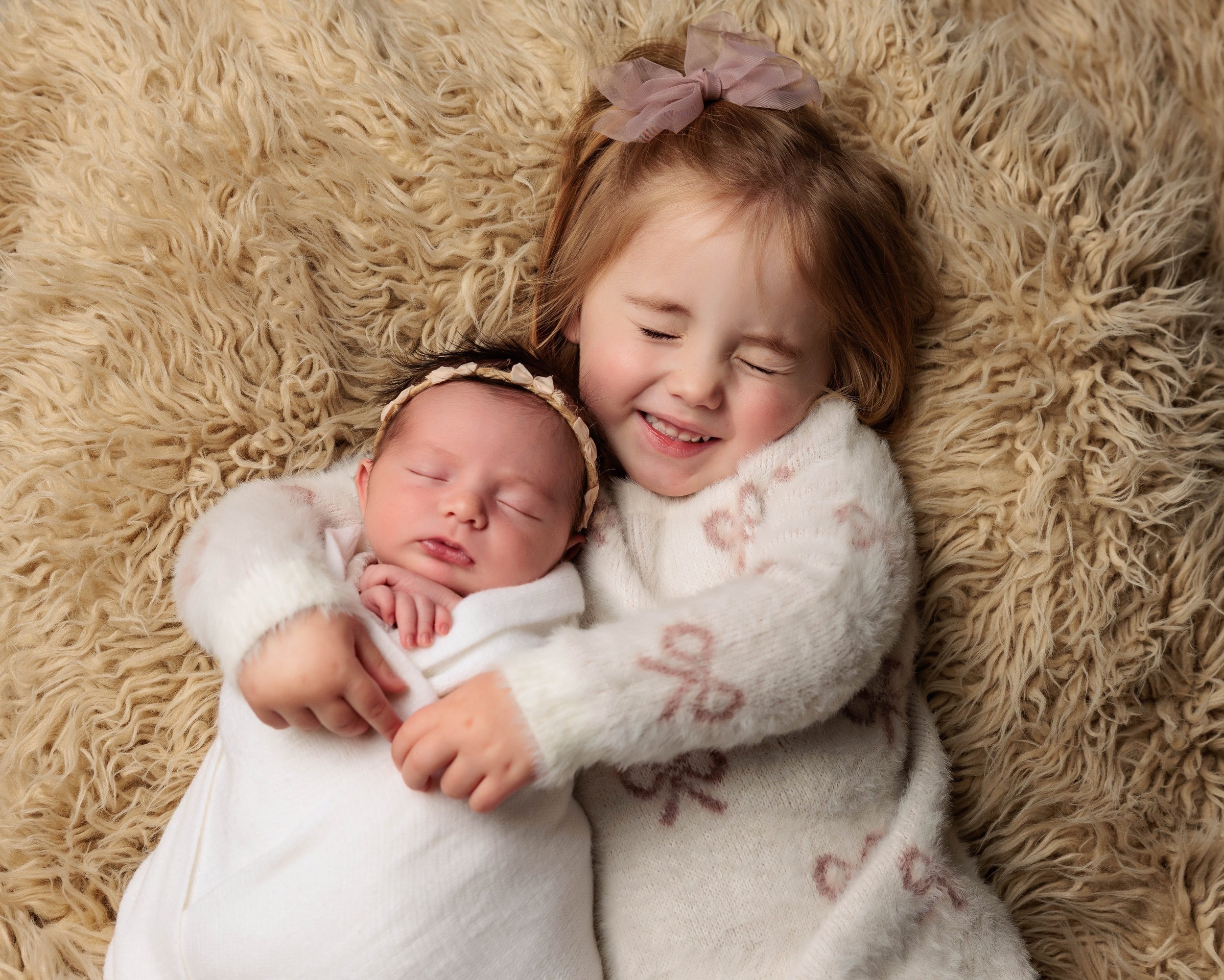 Two young sisters, one a newborn, cuddling on a soft beige furry blanket, both with closed eyes and happy expressions.