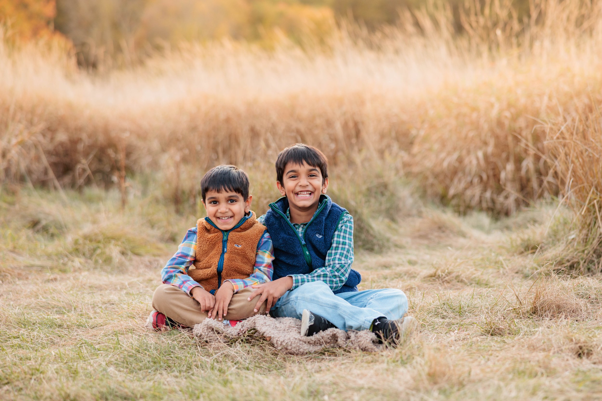 Two young boys sitting on a grassy field, smiling and enjoying nature in an outdoor setting.