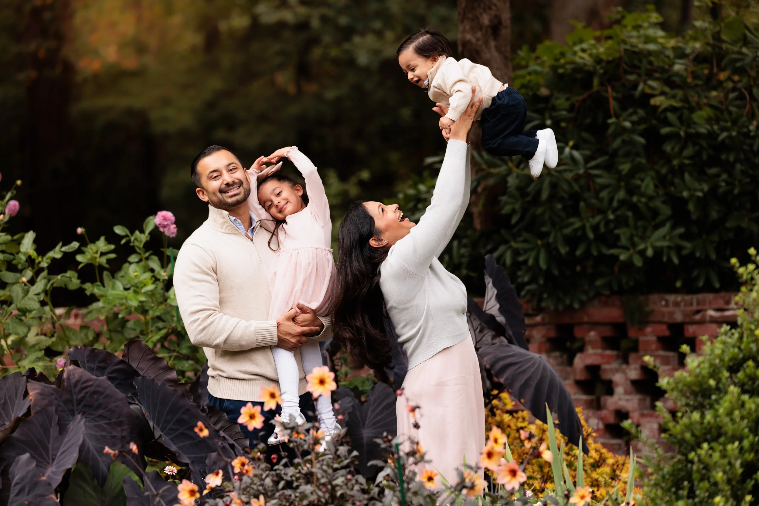 Family of four enjoying time together outdoors among plants, with a woman lifting a young girl into the air.
