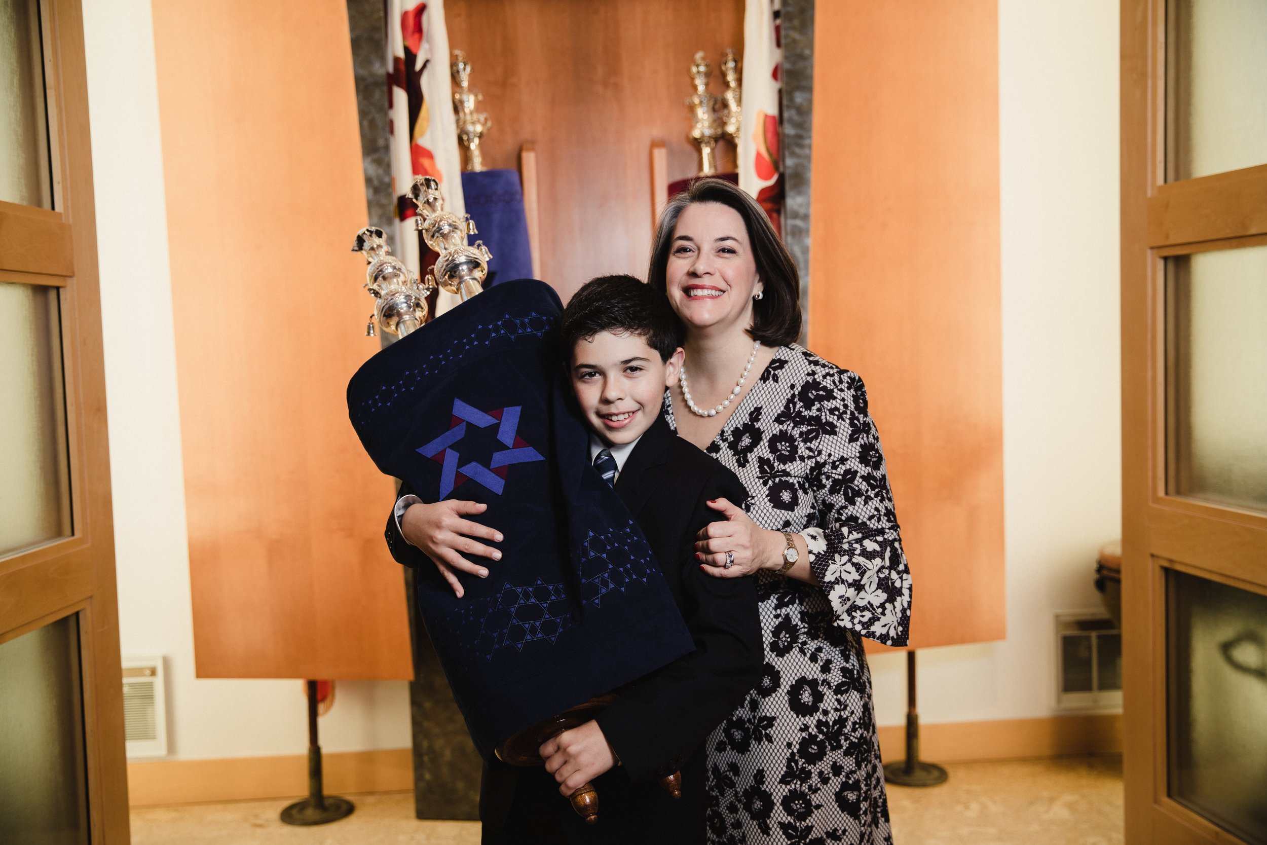 A woman and a boy smiling and hugging, with the boy holding a menorah, in a room decorated with flags and other items in the background.
