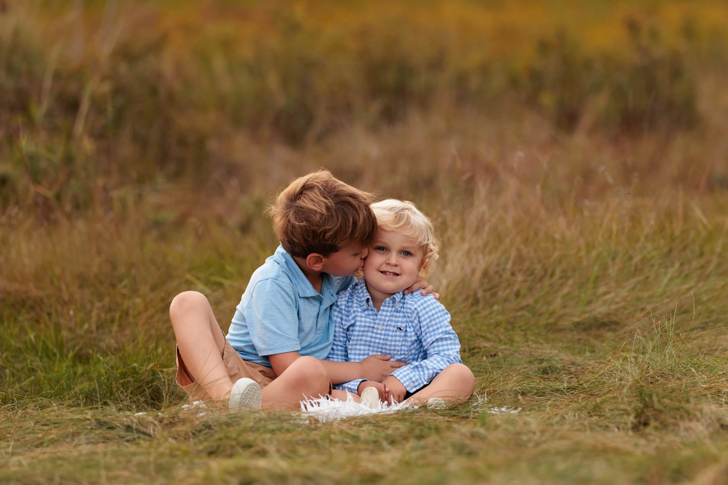 Two young boys sitting on grass in a field, one whispering to the other, who is smiling, with autumn-colored foliage in the background.
