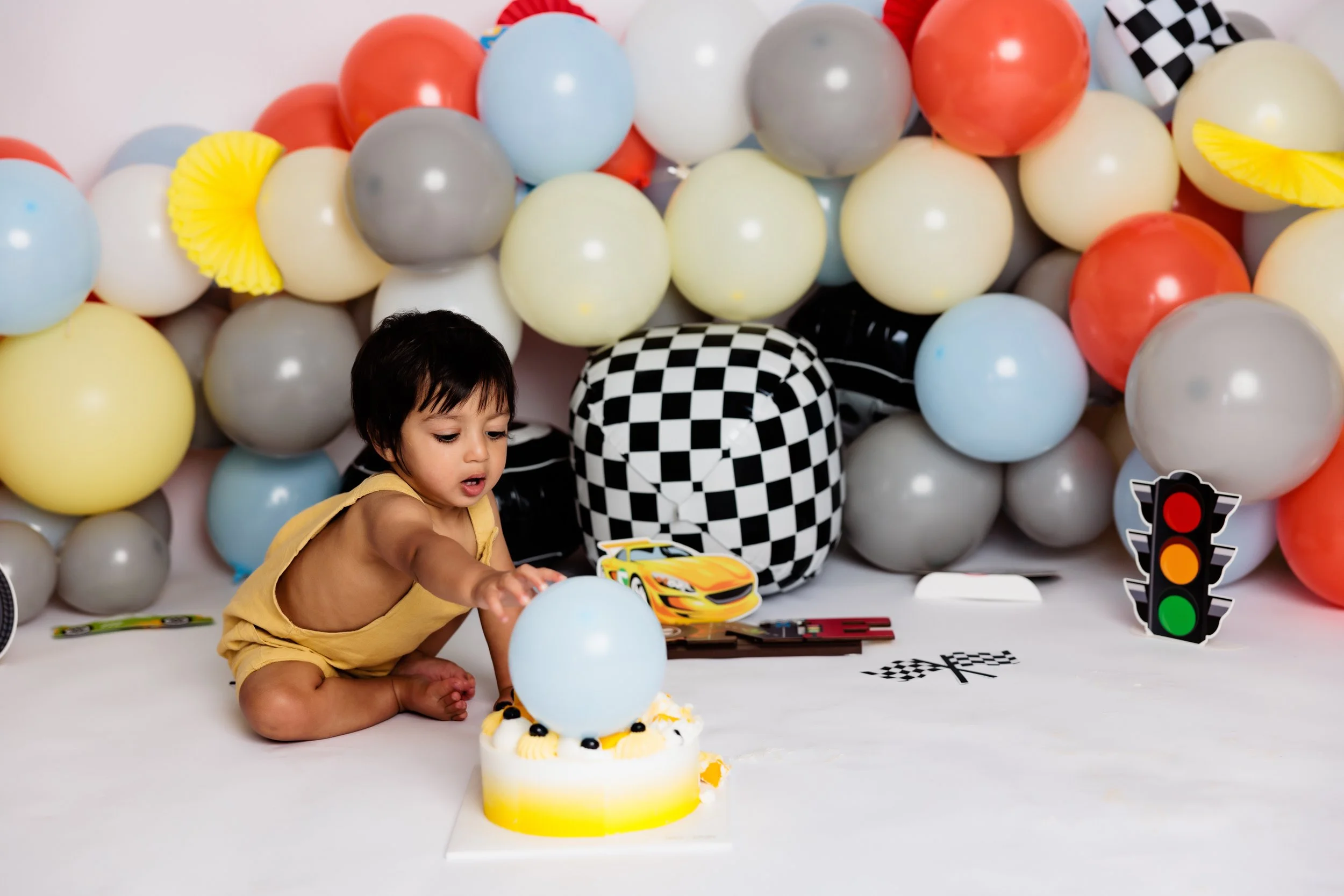 Young child in yellow outfit at birthday celebration with balloons, cake, and racing-themed decorations.