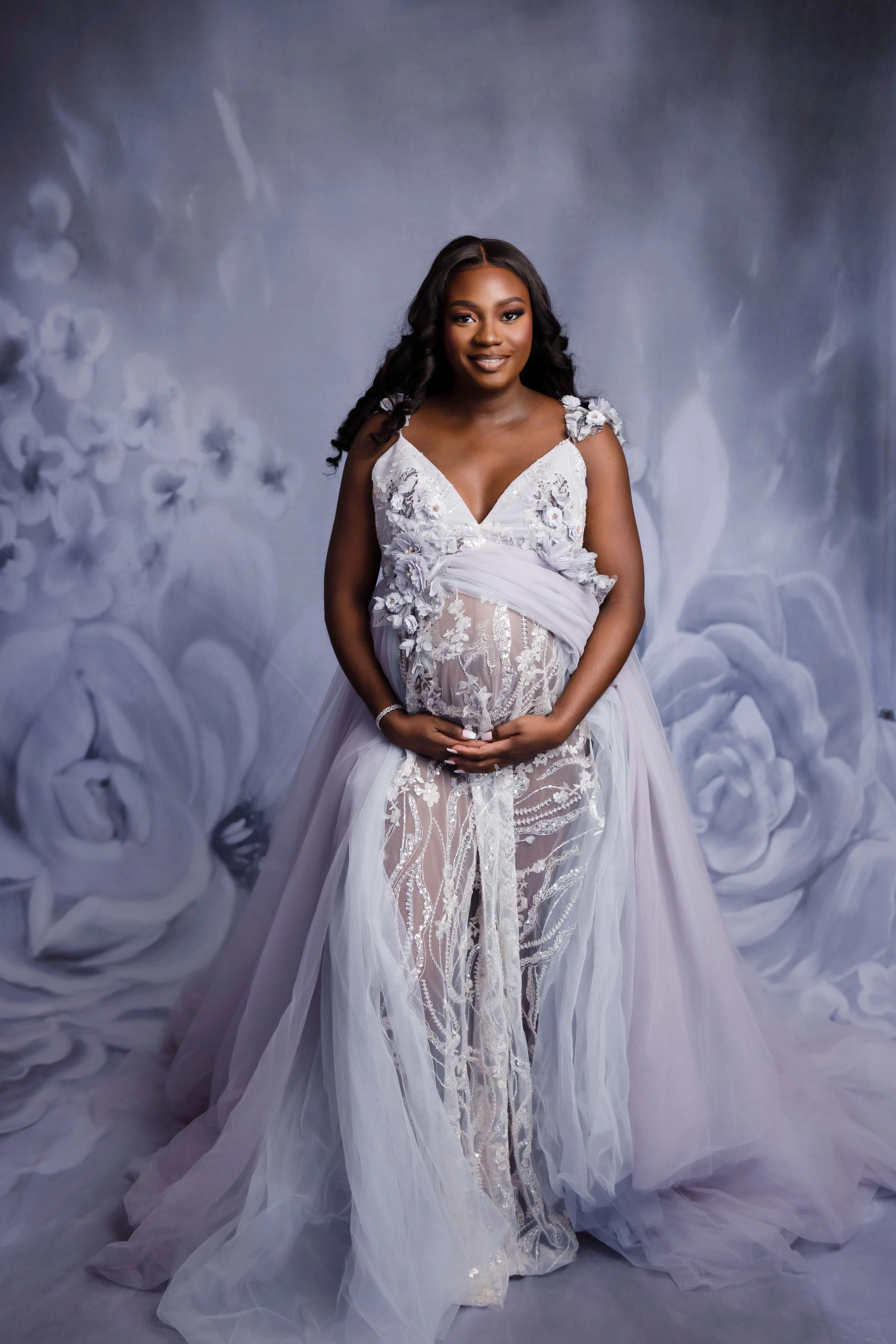 Pregnant woman in a white lace wedding dress standing in front of a floral background.