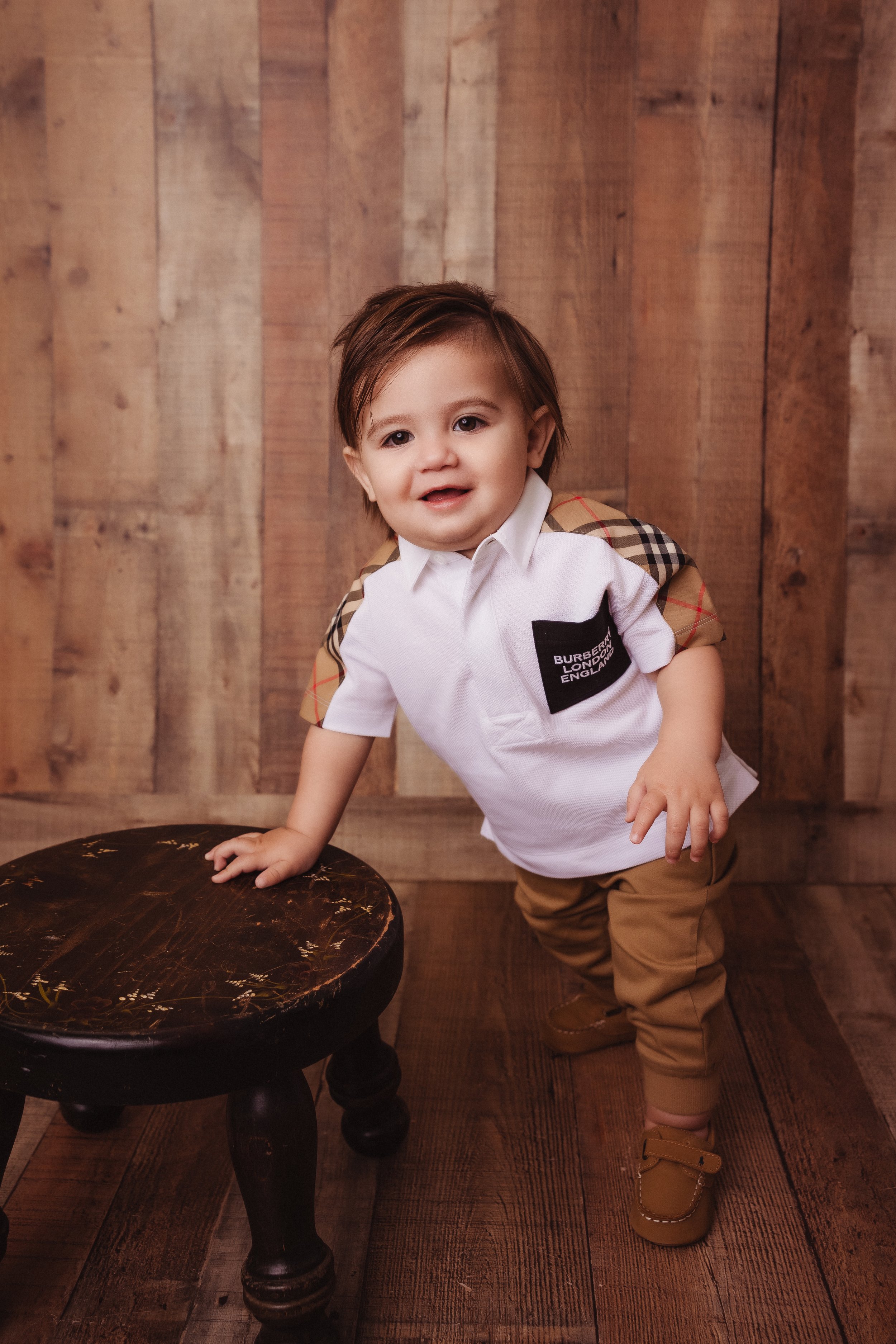 A toddler boy with brown hair, wearing a white shirt with a black pocket, tan pants, and tan shoes, standing next to a small dark wooden stool, against a wooden panel background.