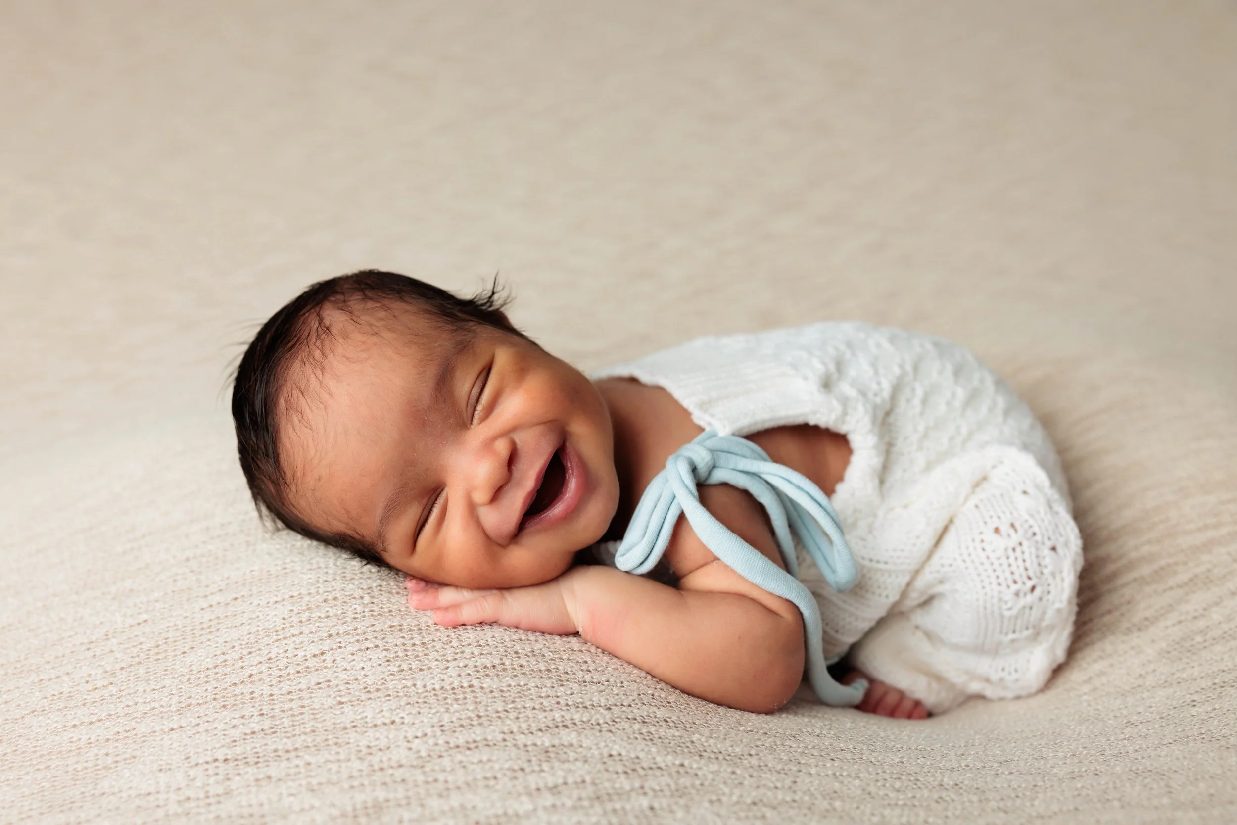 A smiling baby lying on a beige textured surface, wearing a white knitted outfit with a light blue ribbon tied in a bow.