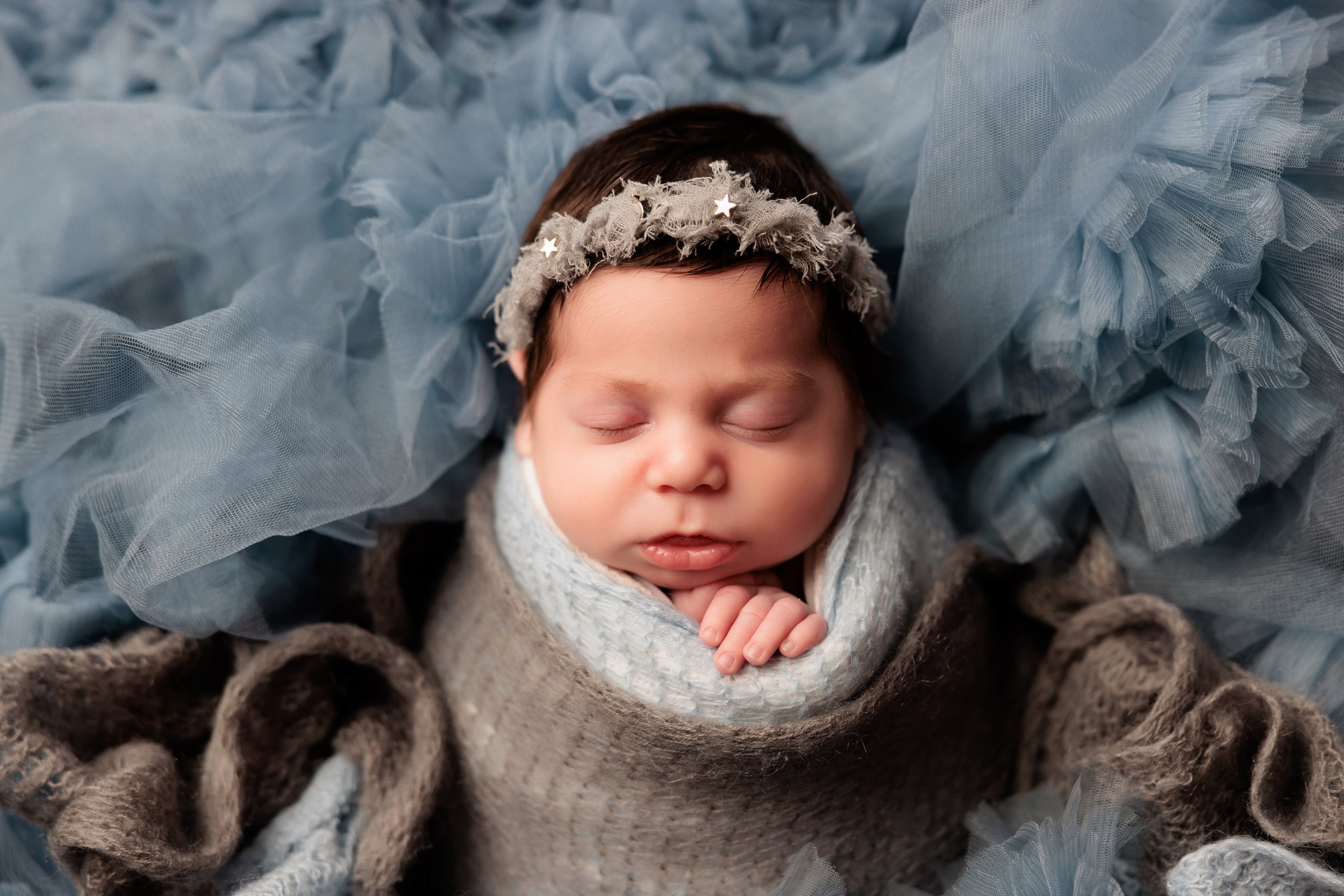 A sleeping baby with a headband, wrapped in a gray knit blanket, surrounded by soft gray tulle fabric.