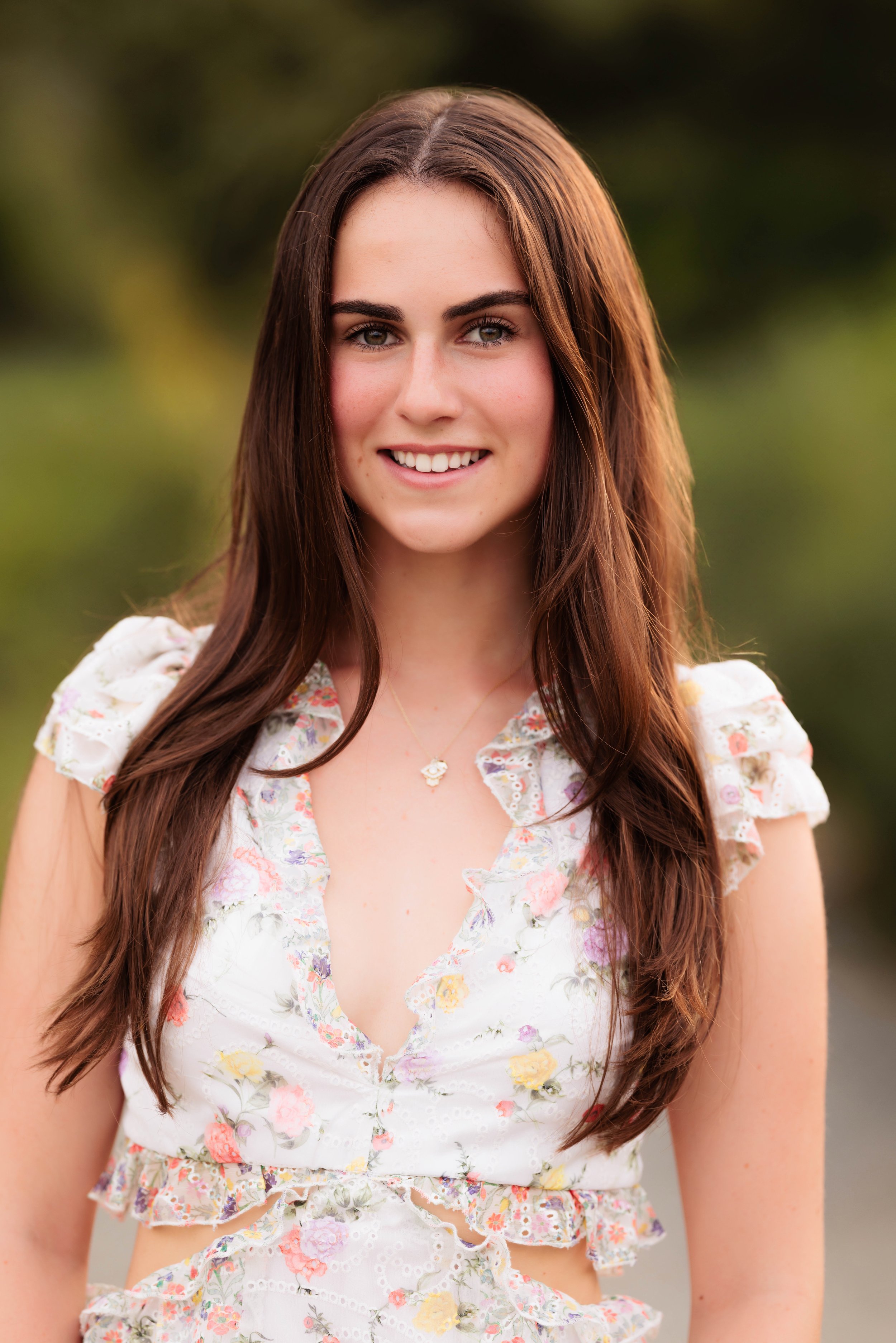 A young woman with long brown hair smiling outdoors, wearing a white floral top with ruffled sleeves and a necklace.