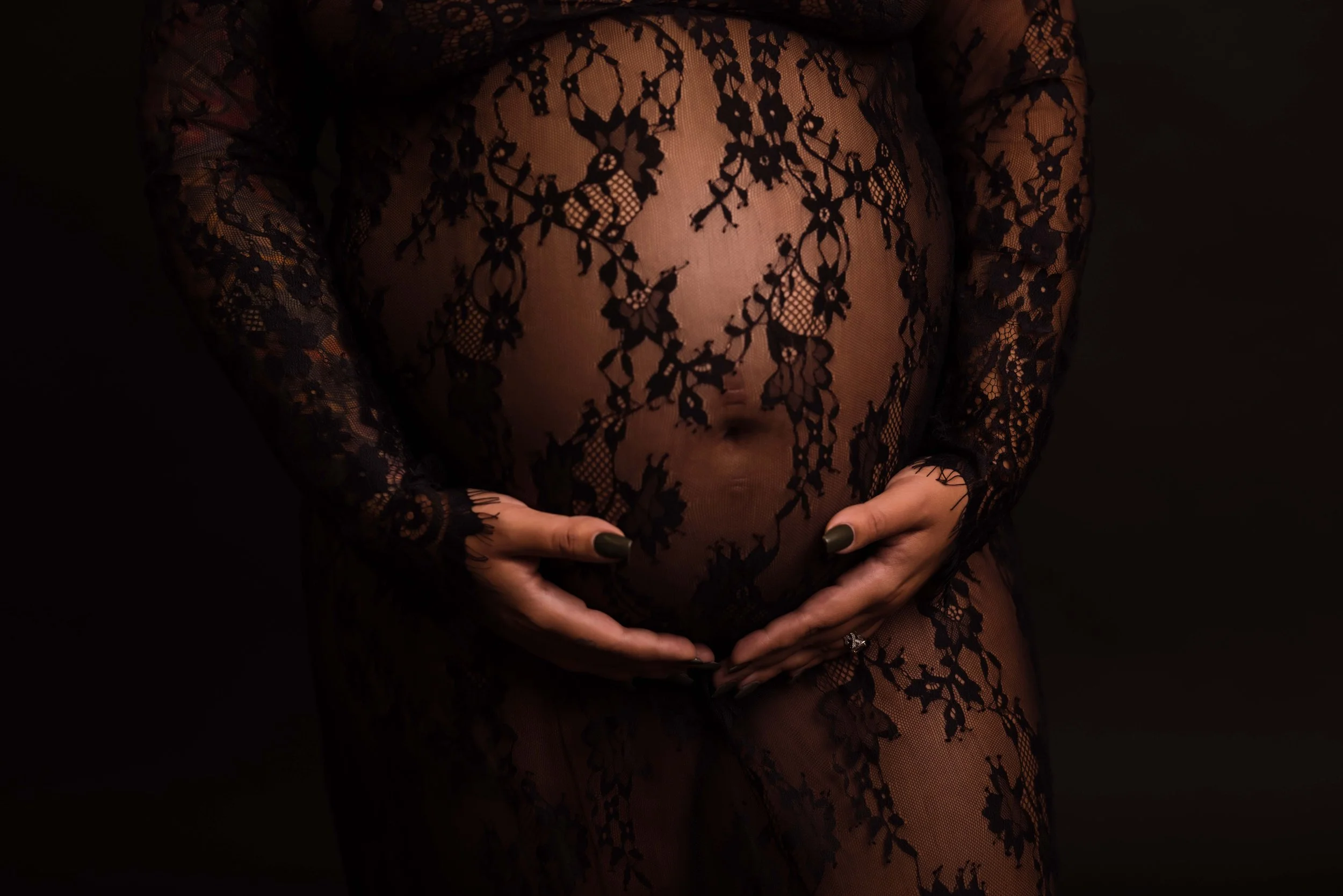 A pregnant woman wearing sheer black lace clothing with floral patterns, gently cradling her belly with both hands.