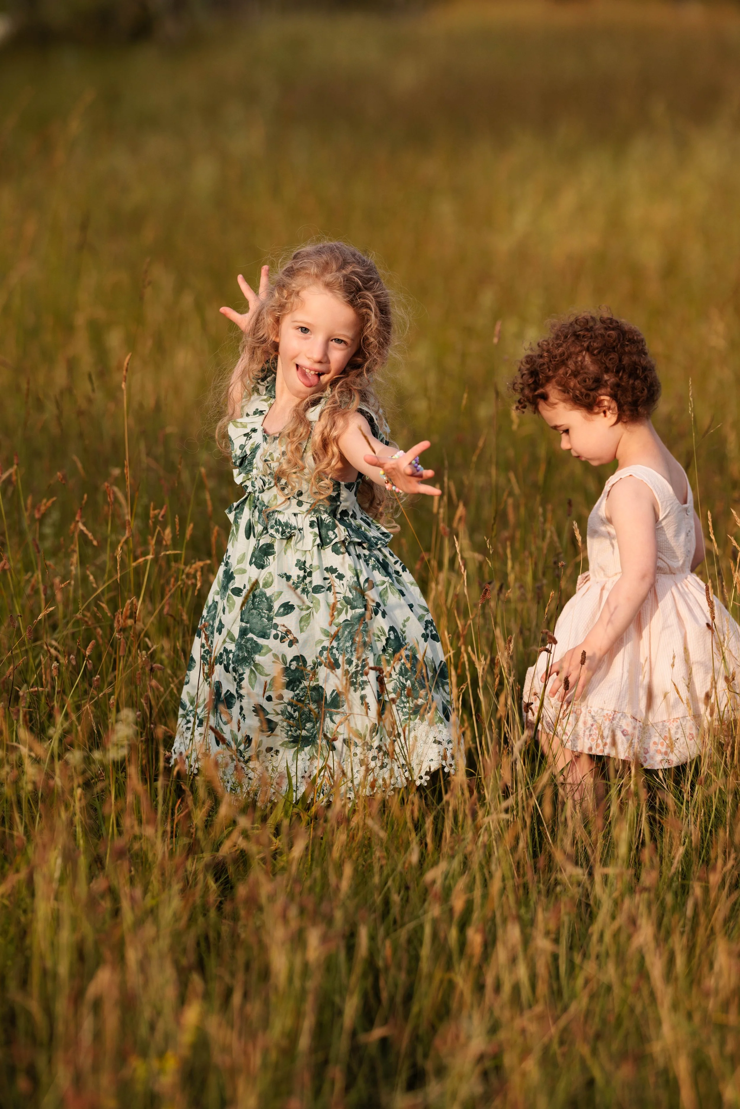 Two young girls in dresses playing and smiling in a grassy field during sunset.