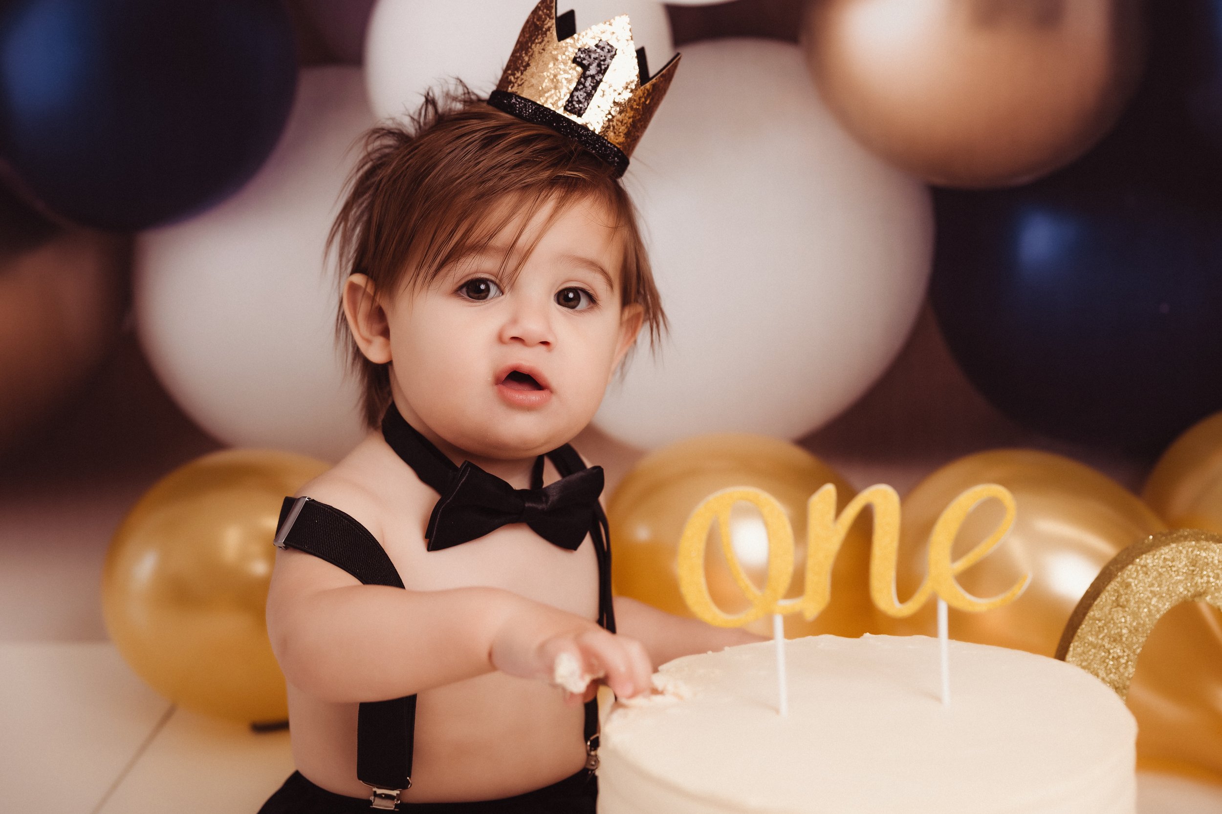 A young child dressed as a birthday boy with a black bow tie and suspenders, wearing a gold "one" crown, celebrating first birthday with a cake and golden balloons in the background.