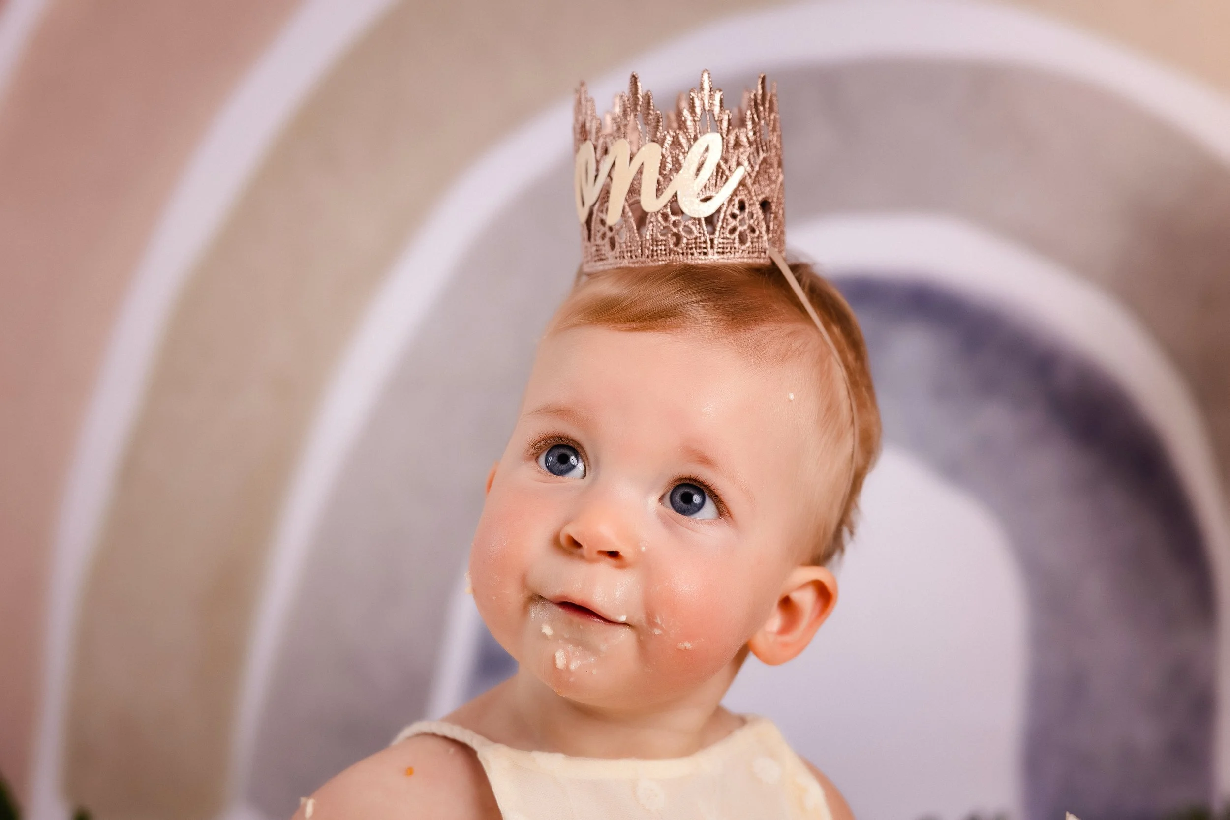 Baby with blue eyes wearing a pink crown with the word 'me' and a cream-colored dress, looking upwards with food on face.