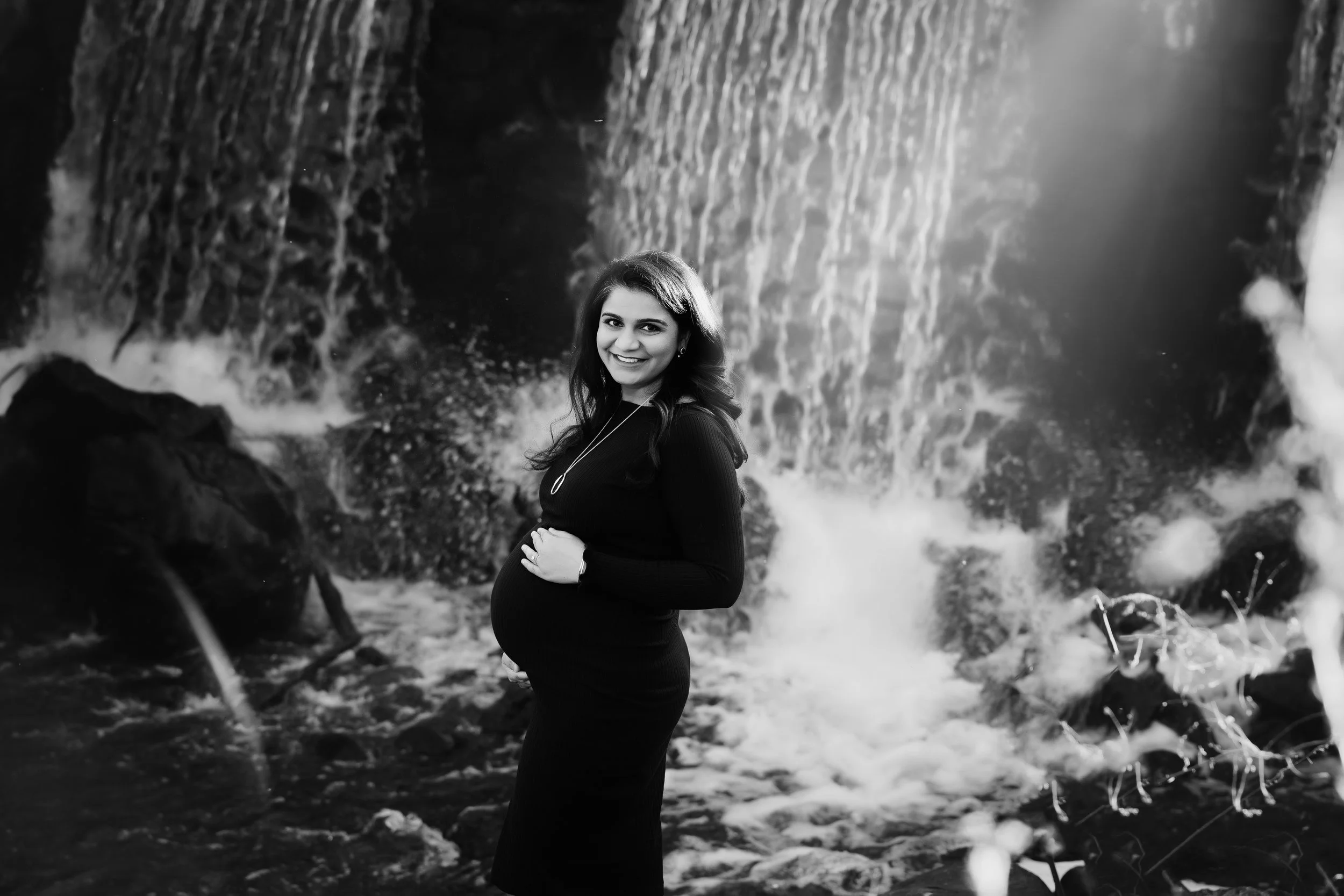 A pregnant woman smiling and holding her belly, standing in front of a waterfall in black and white.