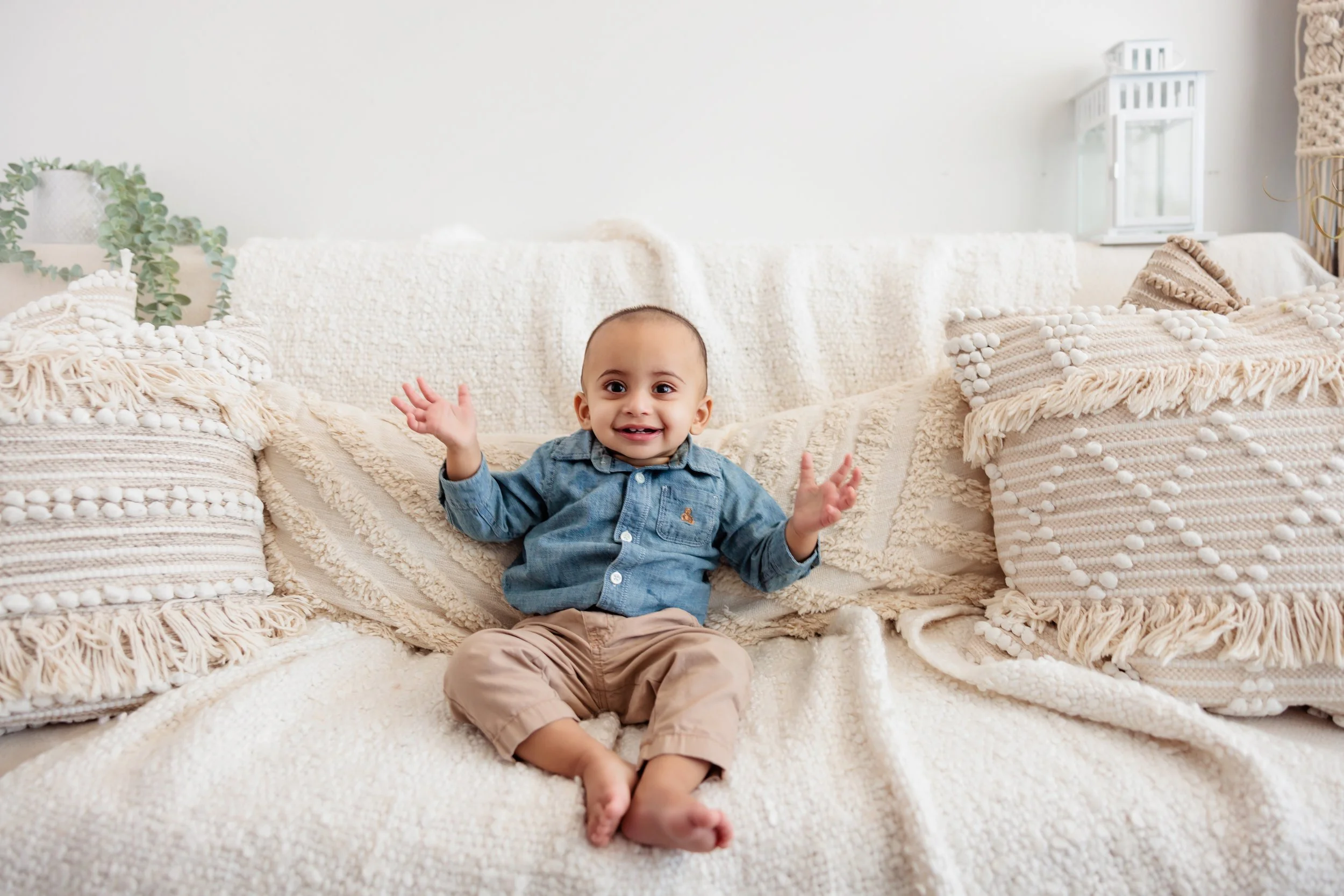 Smiling toddler sitting on a cream-colored textured sofa with decorative pillows, in a cozy living room with a white wall and small plants in the background.