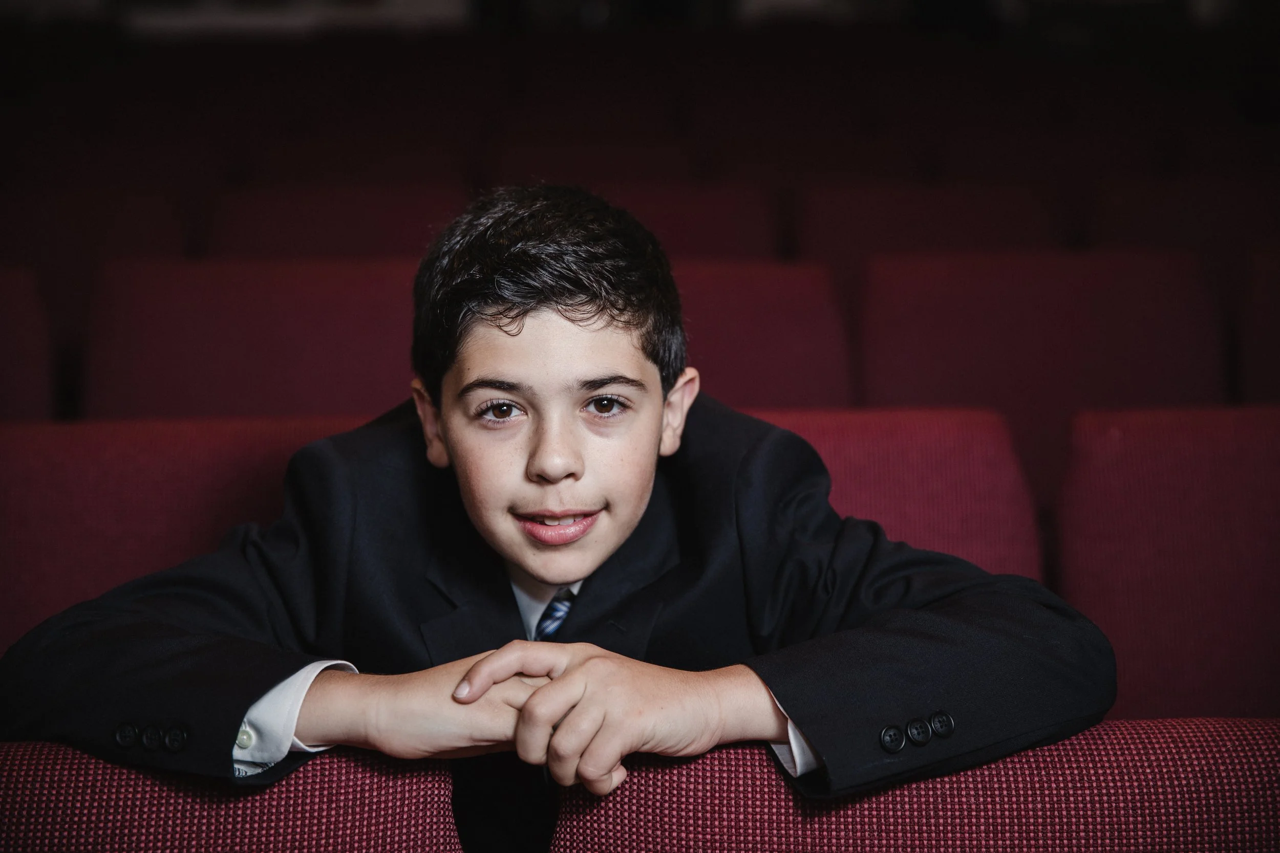 A young boy with short dark hair and light skin, sitting in a theater, dressed in a black suit with a white shirt, leaning forward with his arms on the back of red theater seat, looking at the camera.