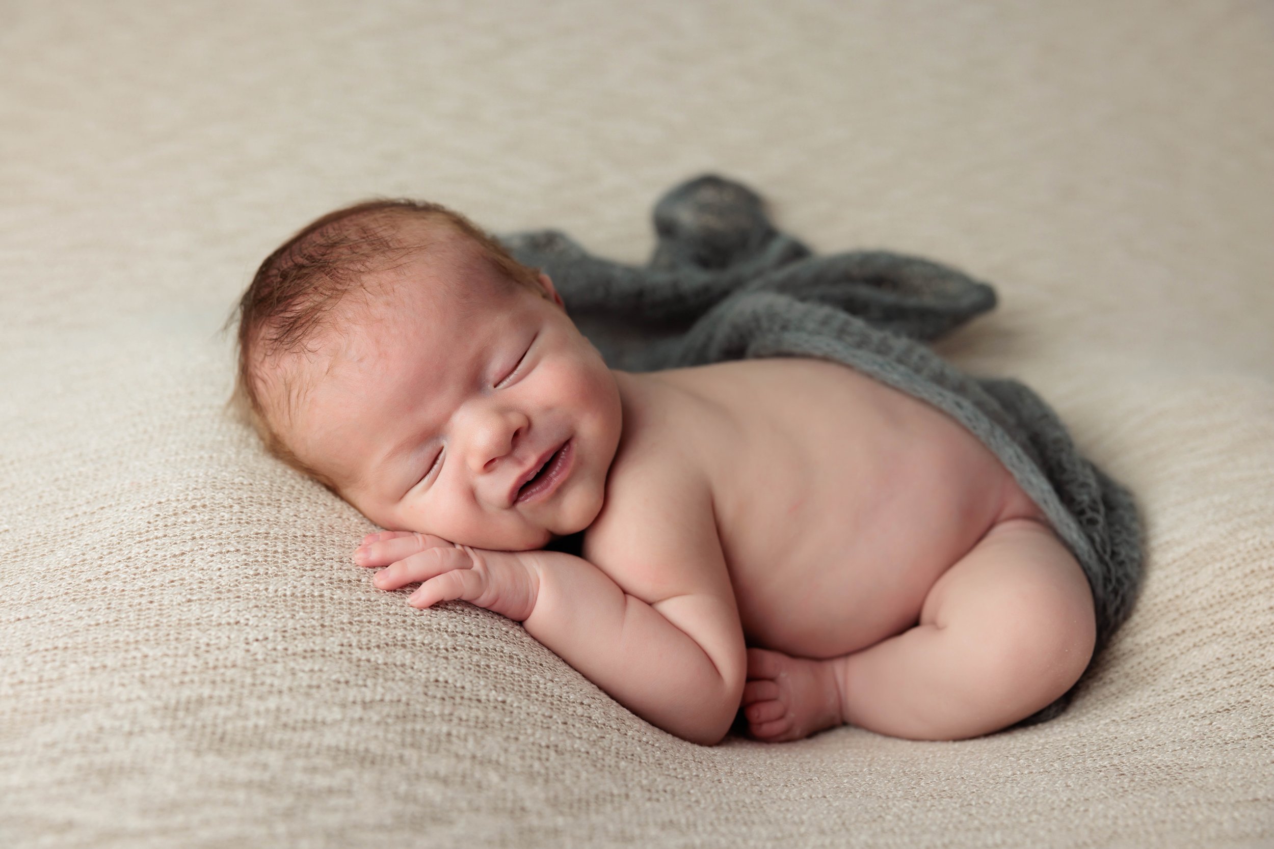 A smiling newborn baby with brown hair, lying on its side on a beige textured surface, wearing a gray knitted tail and ears blanket.