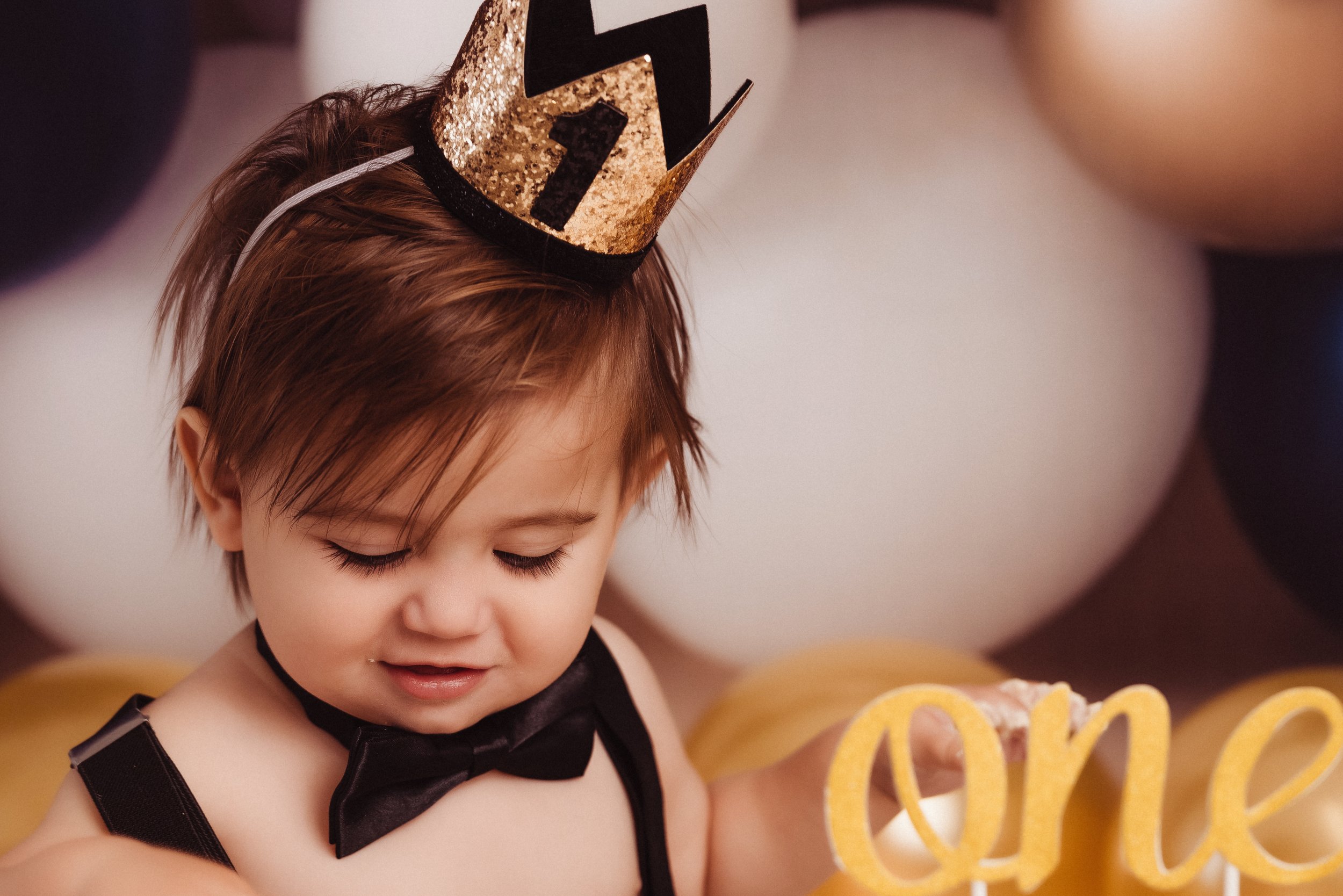 A young child with short brown hair wearing a gold glittery birthday crown, a black bow tie, and a black strap top, holding a yellow decorative cake topper that spells 'one'.