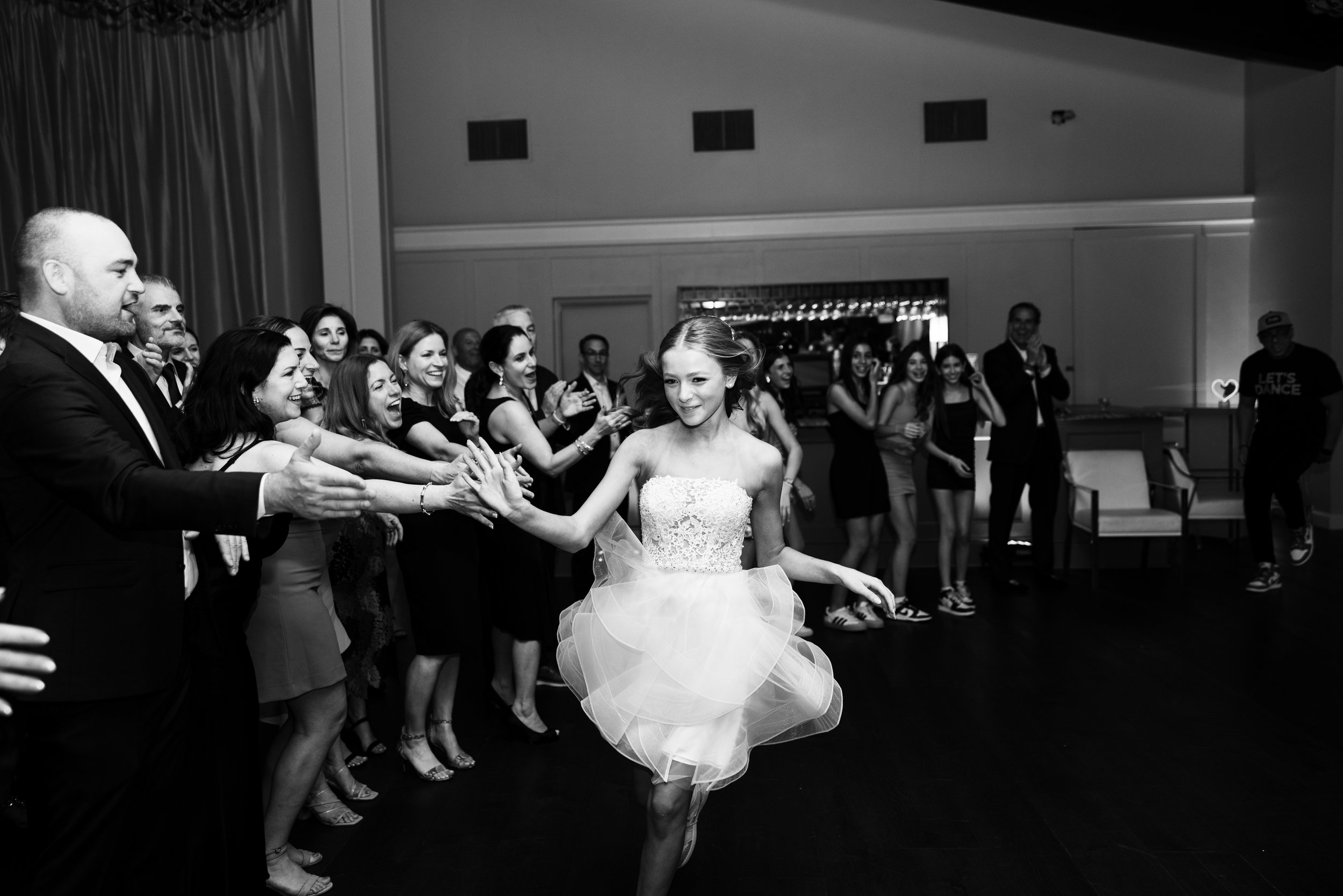 A young girl in a white wedding dress dancing with a group of people at a celebration, with some clapping and cheering.