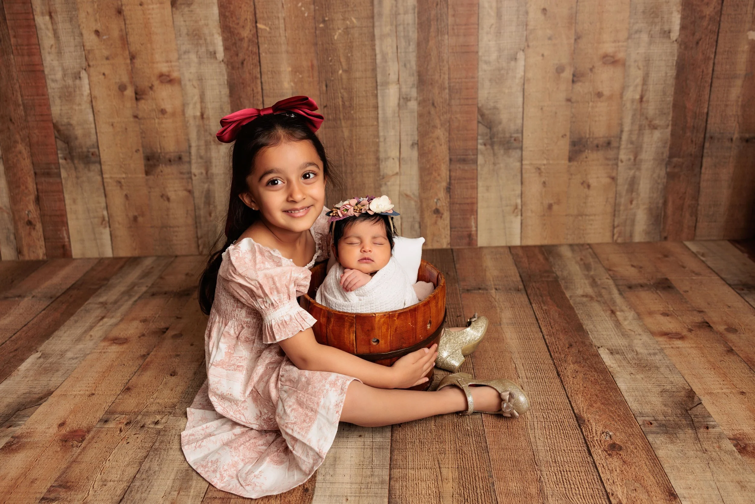 A young girl in a pink dress with bow headband sitting on wooden floor, holding a sleeping baby girl in a wooden bowl, both against a wooden backdrop.