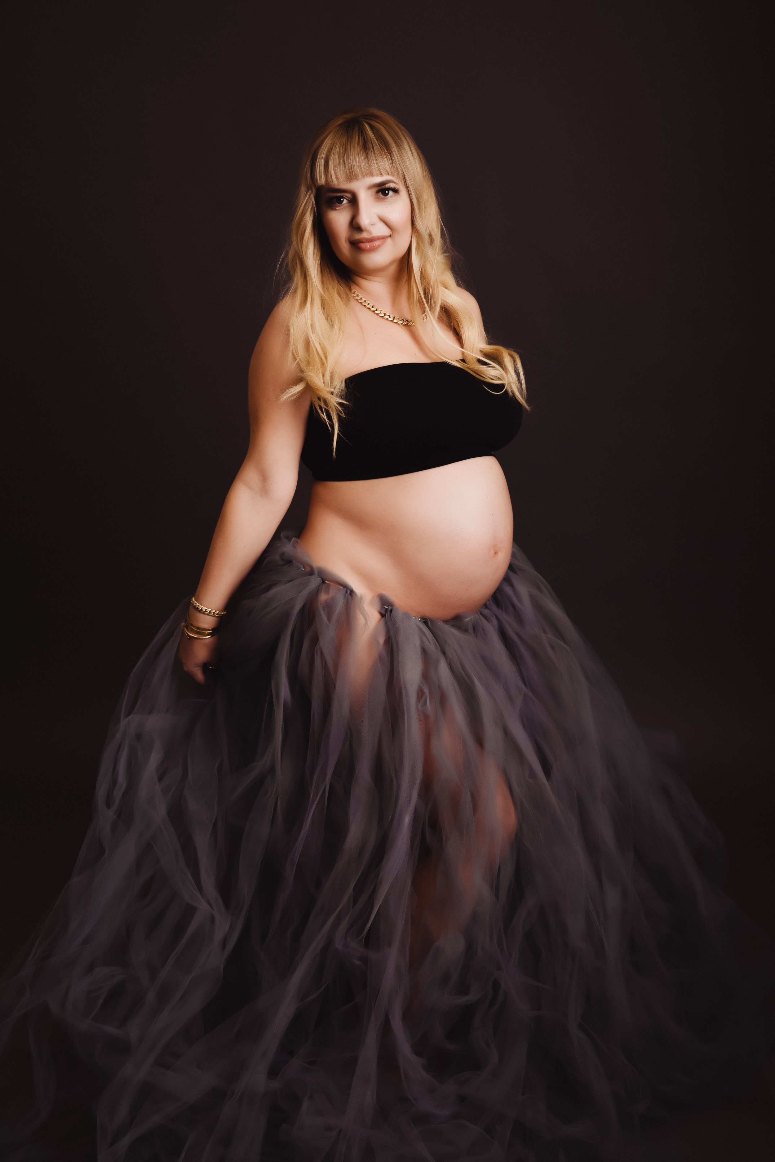 Pregnant woman wearing a black strapless top and a tulle skirt, standing against a dark background.