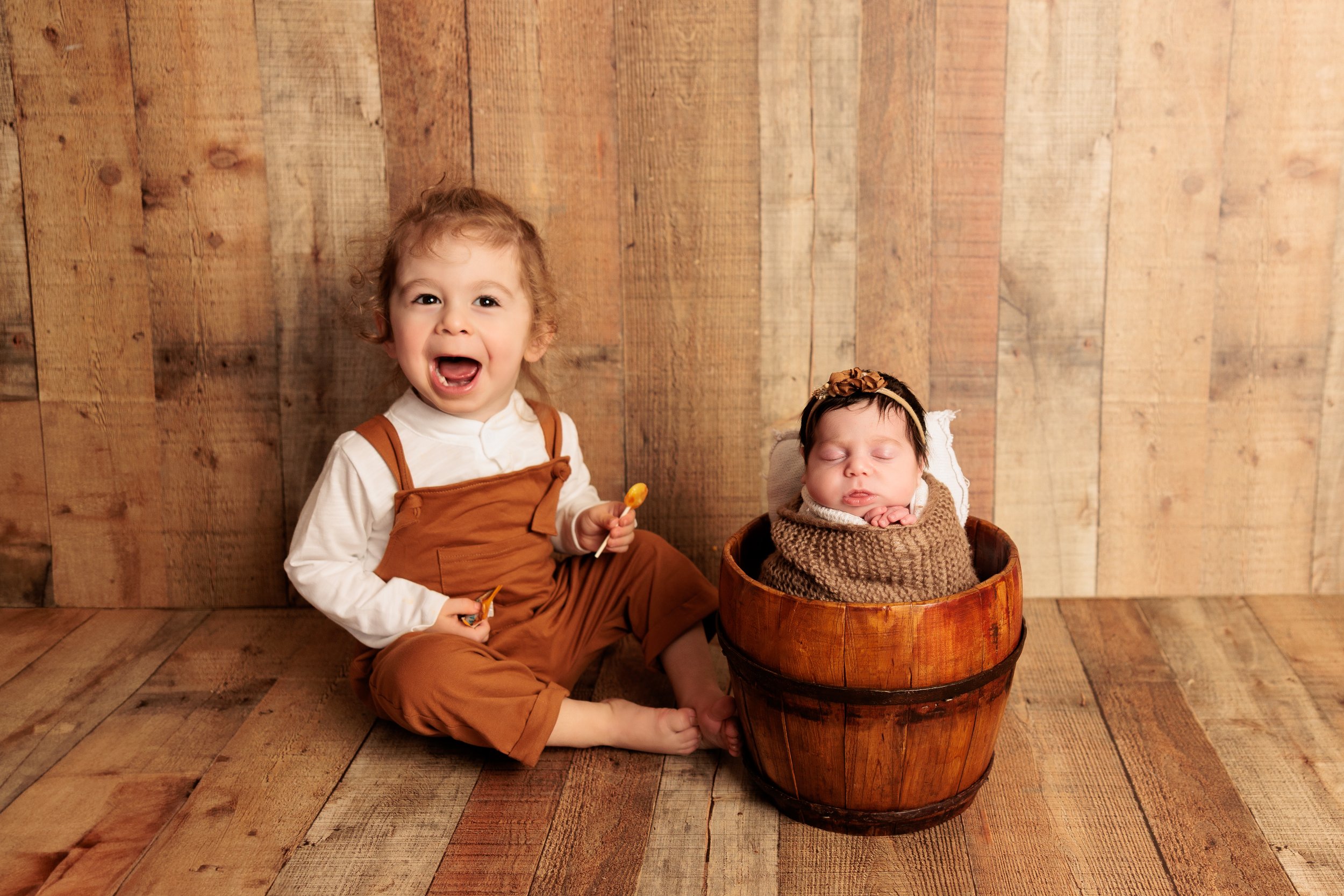 A young child with curly hair, wearing a white shirt and brown overalls, sitting on a wooden floor next to a sleeping baby wrapped in brown knit and in a wooden bucket. The child is smiling widely and holding a lollipop.