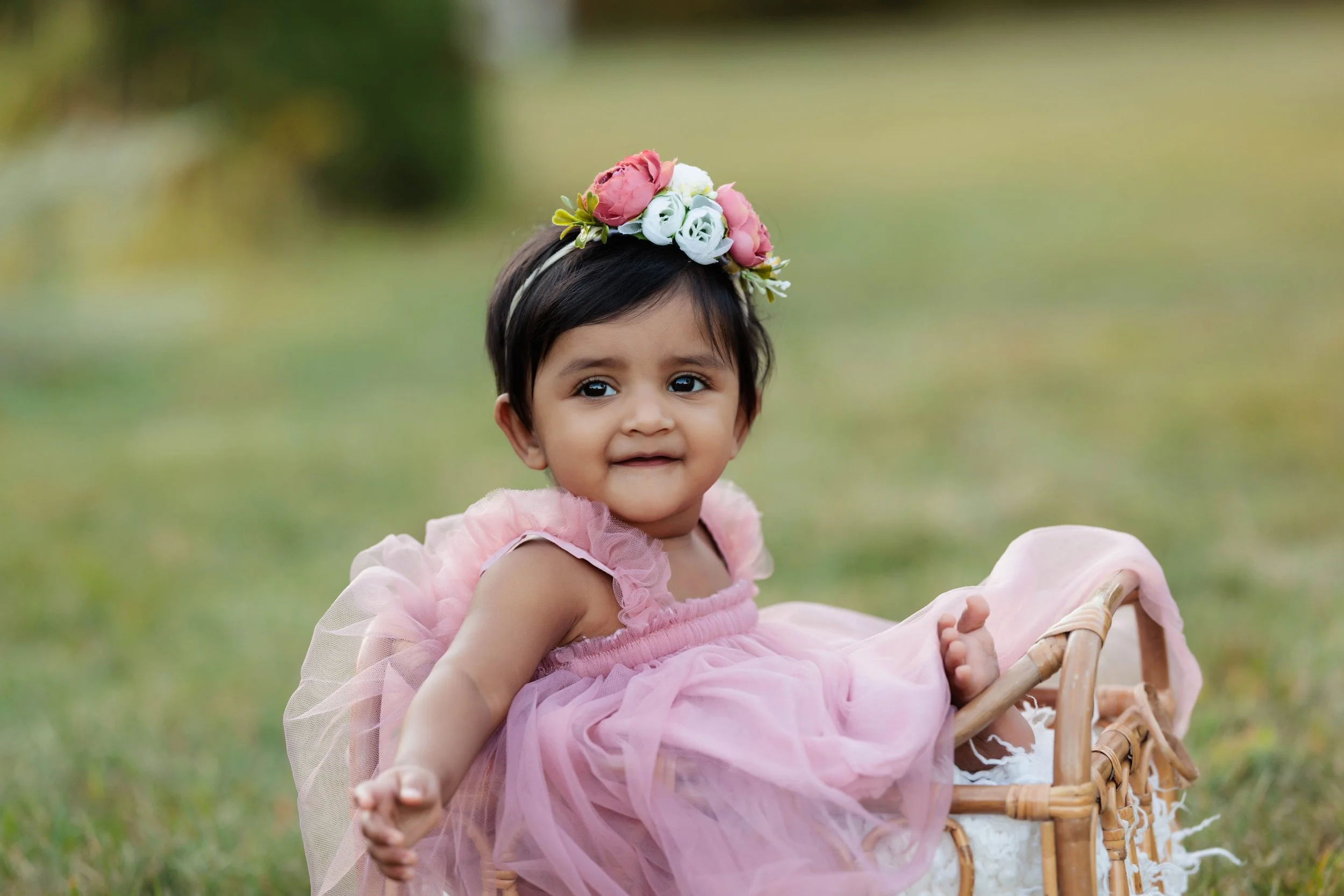 A young girl with short black hair, wearing a pink dress with ruffled sleeves, and a flower crown on her head, sitting in a small wicker basket outdoors on green grass.