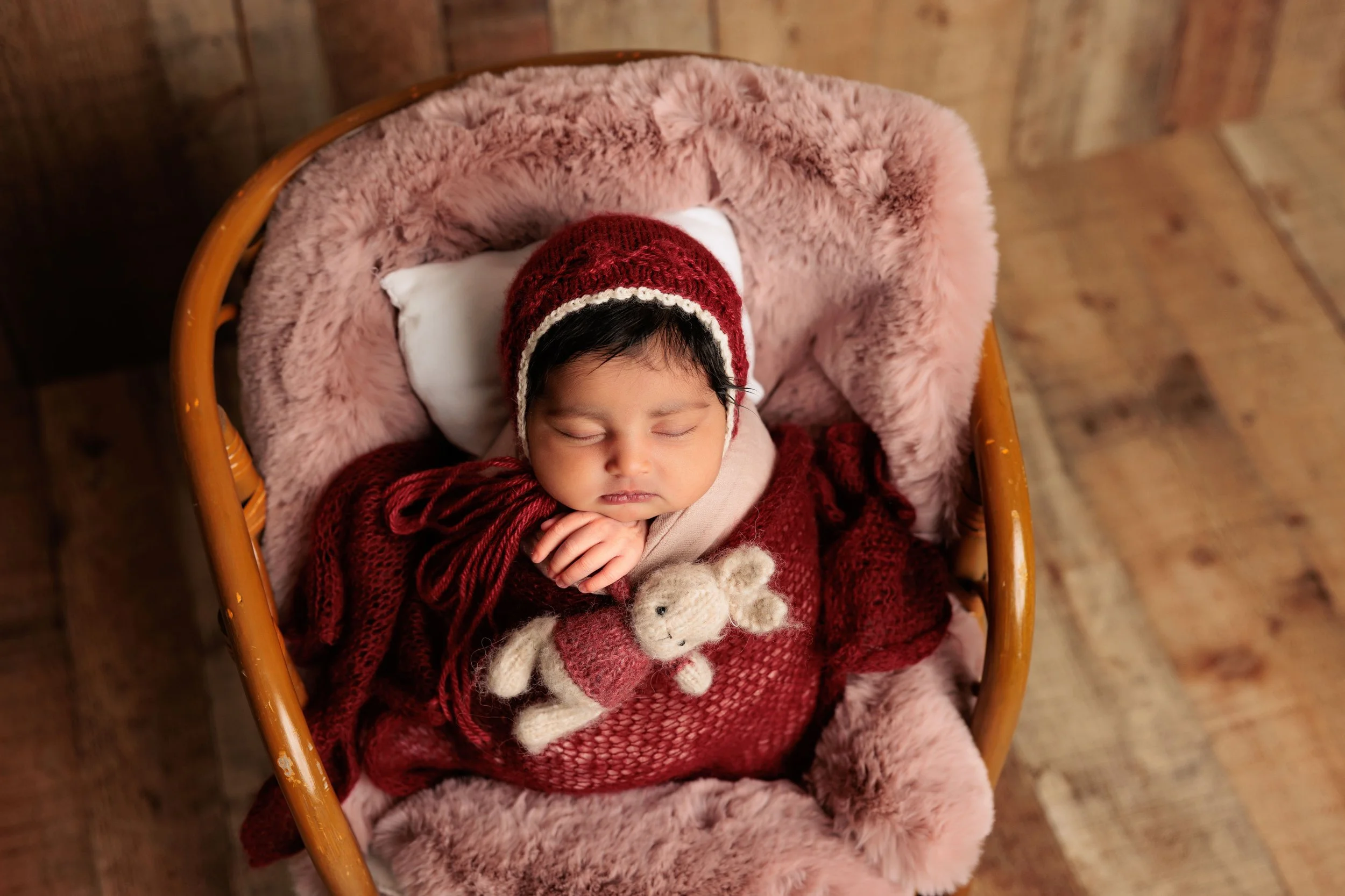 Young baby sleeping in a cozy pink fuzzy blanket in a cradle, wearing a red knitted hat, with a small stuffed bunny toy.