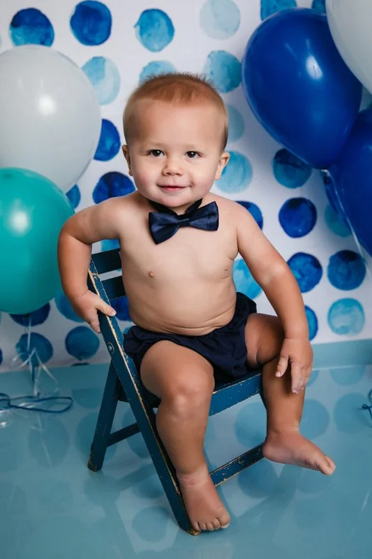 A smiling young child sitting shirtless on a small blue chair, wearing a black bow tie and black shorts, with colorful balloons and a polka dot background in blue and white behind him.