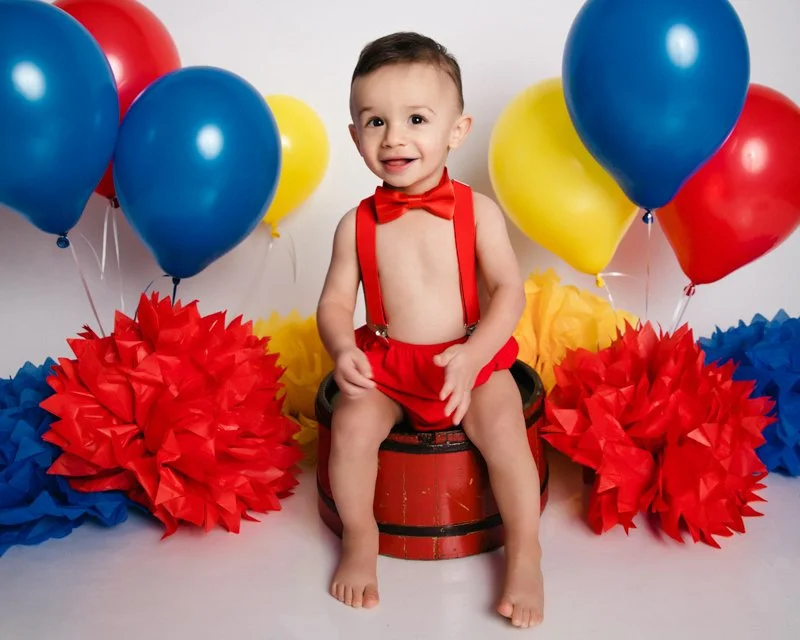 A smiling baby boy dressed in red suspenders and a red bow tie sitting on a barrel surrounded by colorful balloons and large tissue paper pom-poms in red, yellow, and blue.