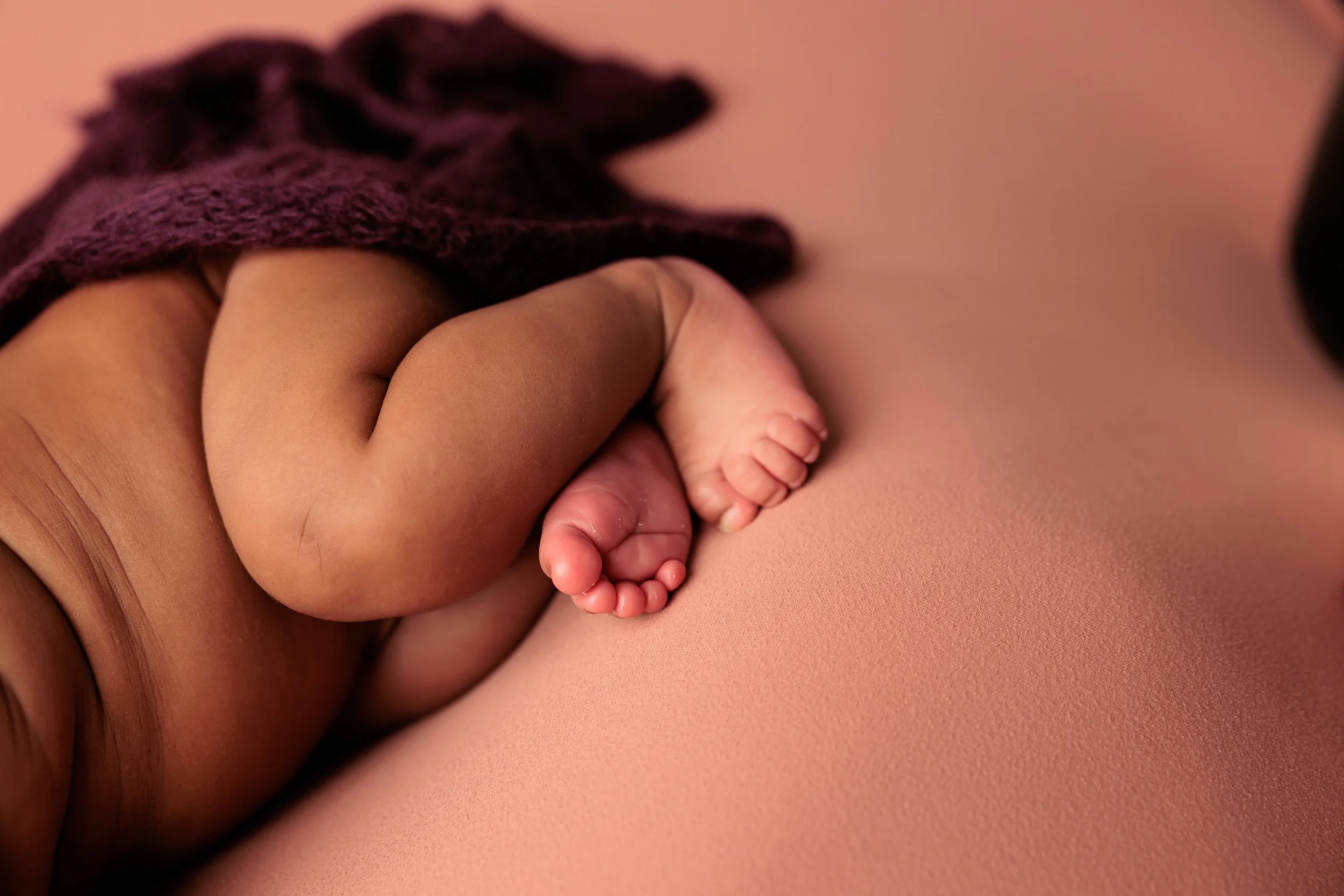 Close-up of a newborn baby's tiny feet and legs, lying on a pink surface with a purple cloth partially covering them.