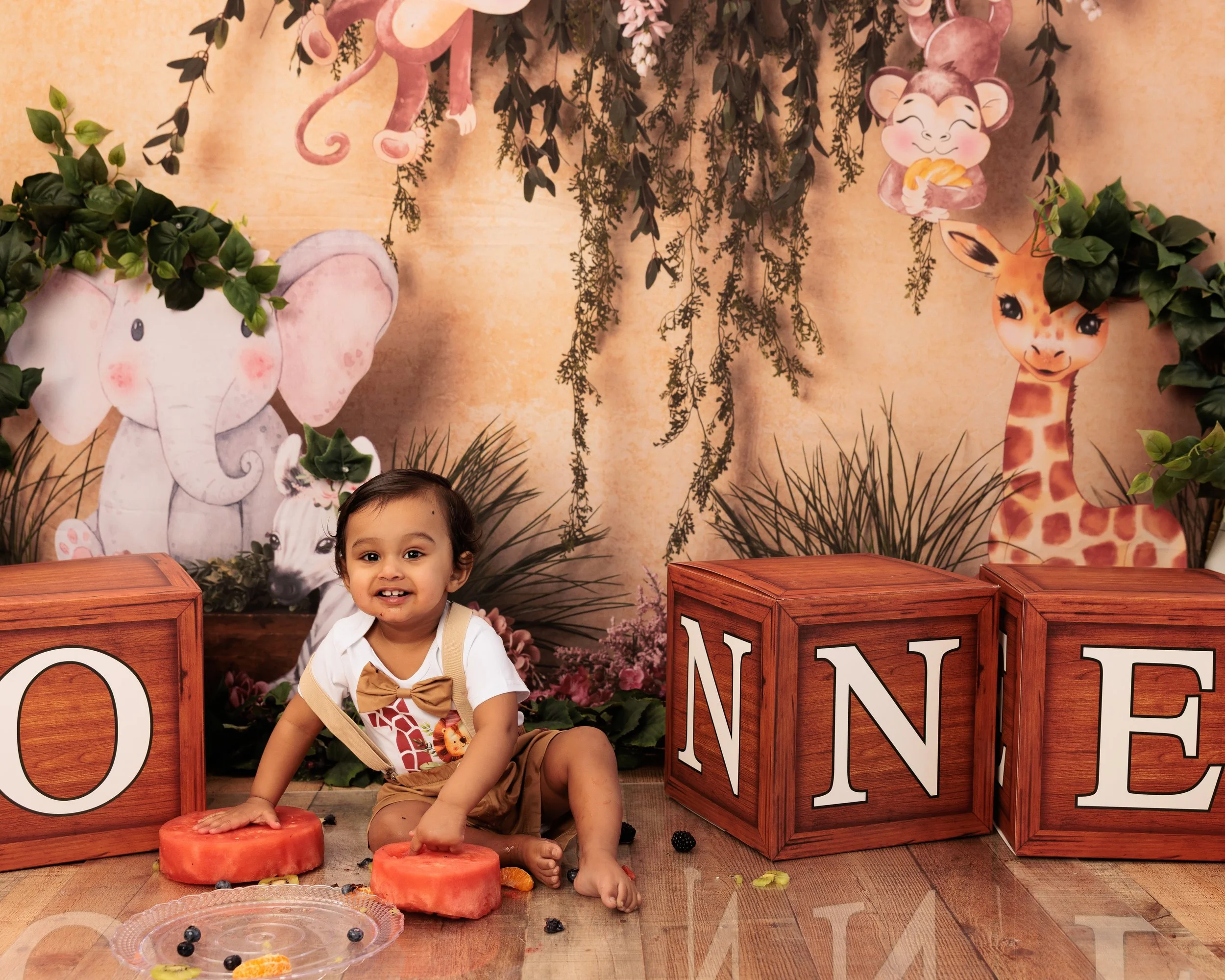 A smiling young girl sitting on a wooden floor, surrounded by large alphabet blocks spelling 'ONE', with a decorated jungle scene backdrop featuring cartoon animals including a giraffe, elephant, tiger, and monkeys, along with greenery and hanging vi