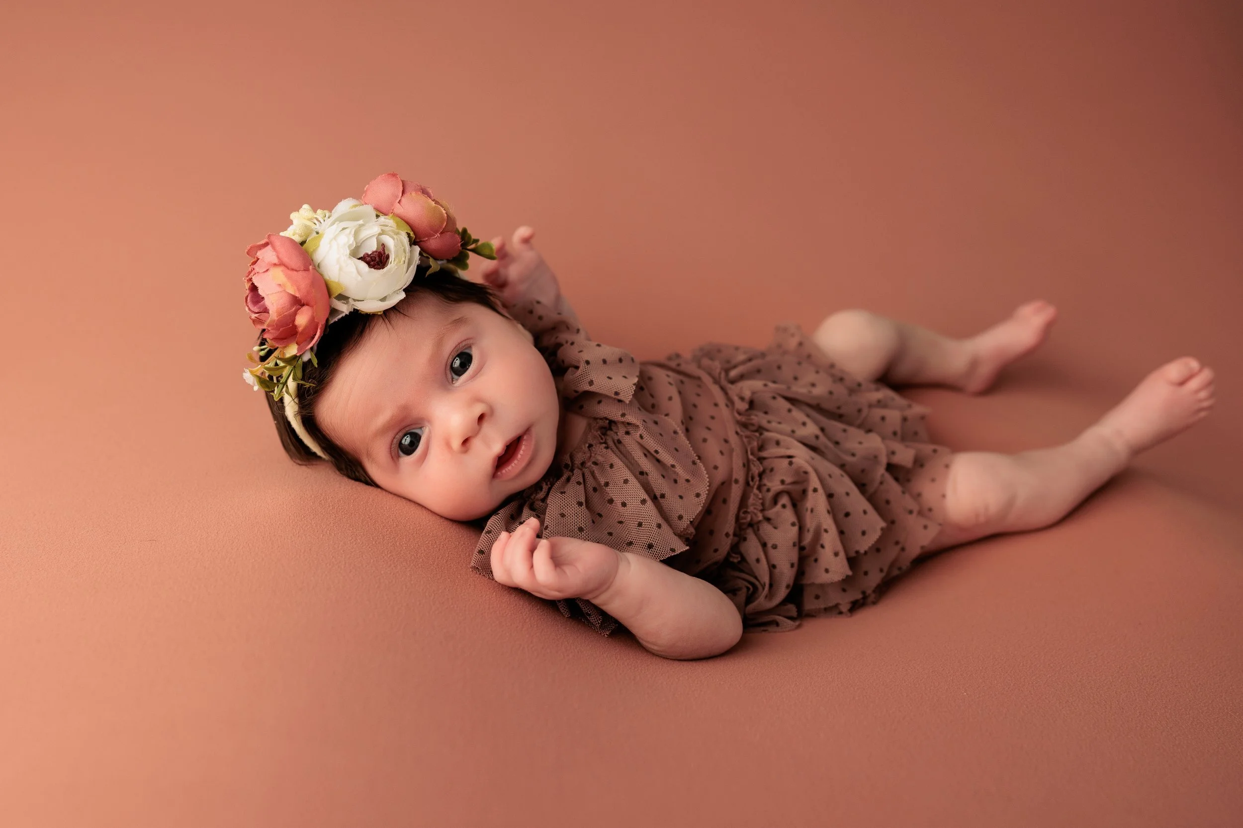 A baby girl lying on a pink surface, wearing a brown polka dot dress and a floral headband with pink and white flowers, looking at the camera.