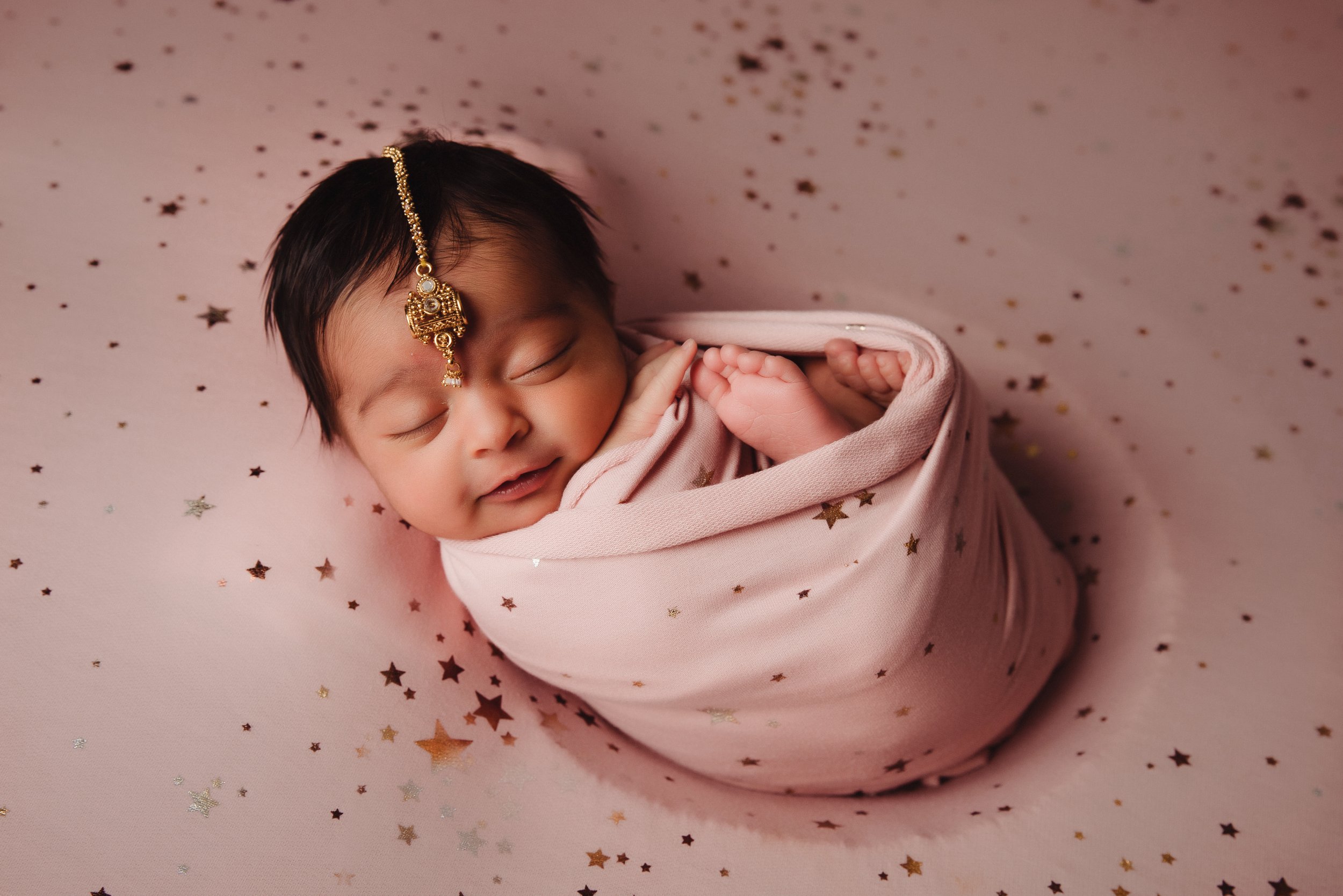 Smiling sleeping baby wrapped in pink blanket with star patterns, wearing a gold headband, lying on star-patterned surface.