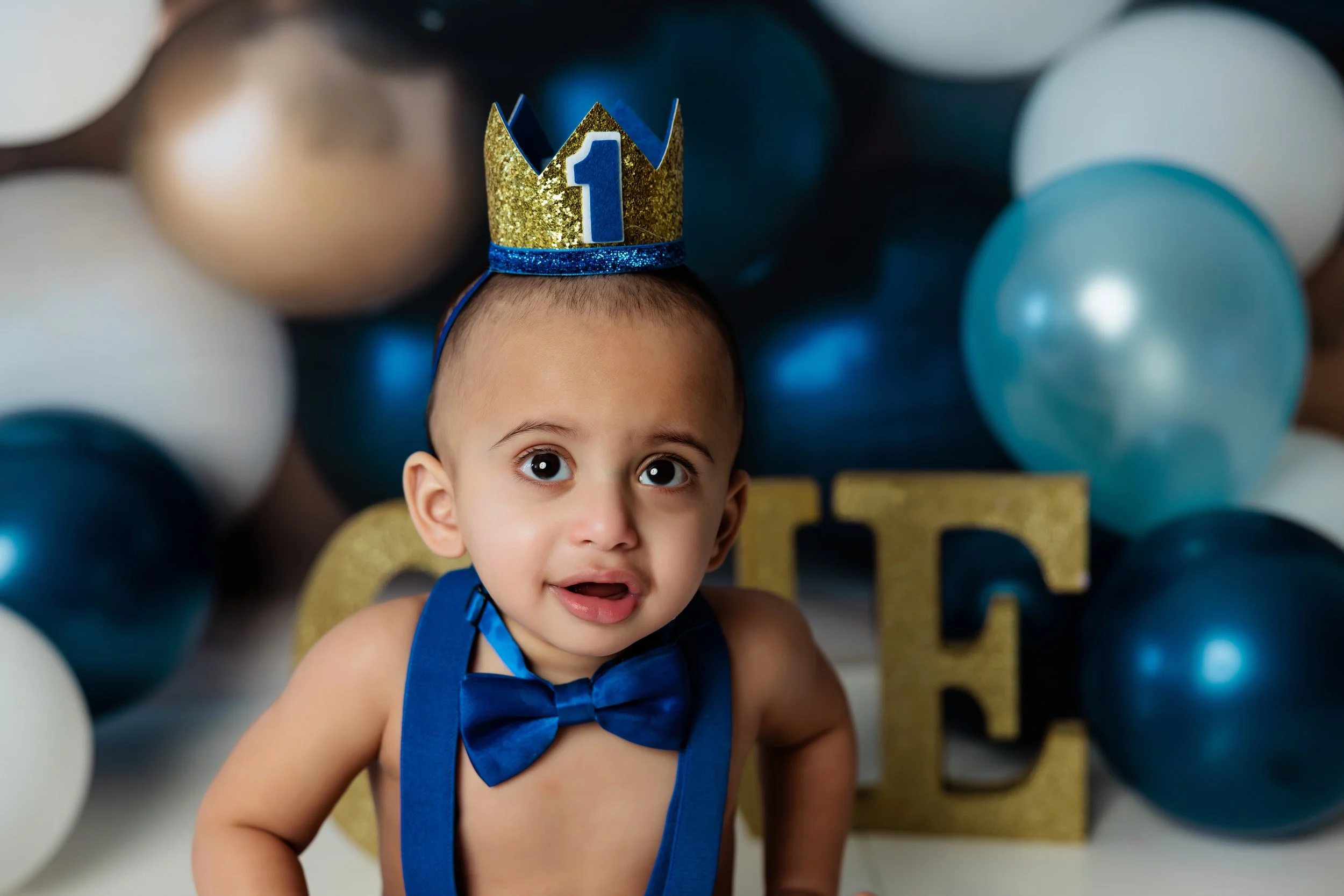A young boy wearing a blue bow tie and a birthday crown with the number 1 stands in front of a birthday party backdrop with balloons and gold decorative letters.